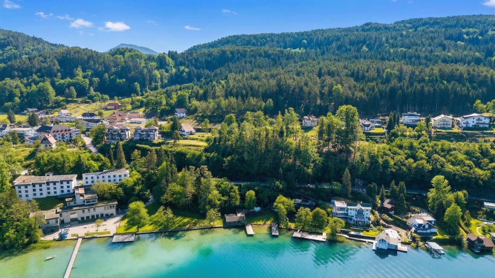 Malerische Luftaufnahme des Sees mit Uferbebauung und bewaldeten Bergen unter blauem Himmel.