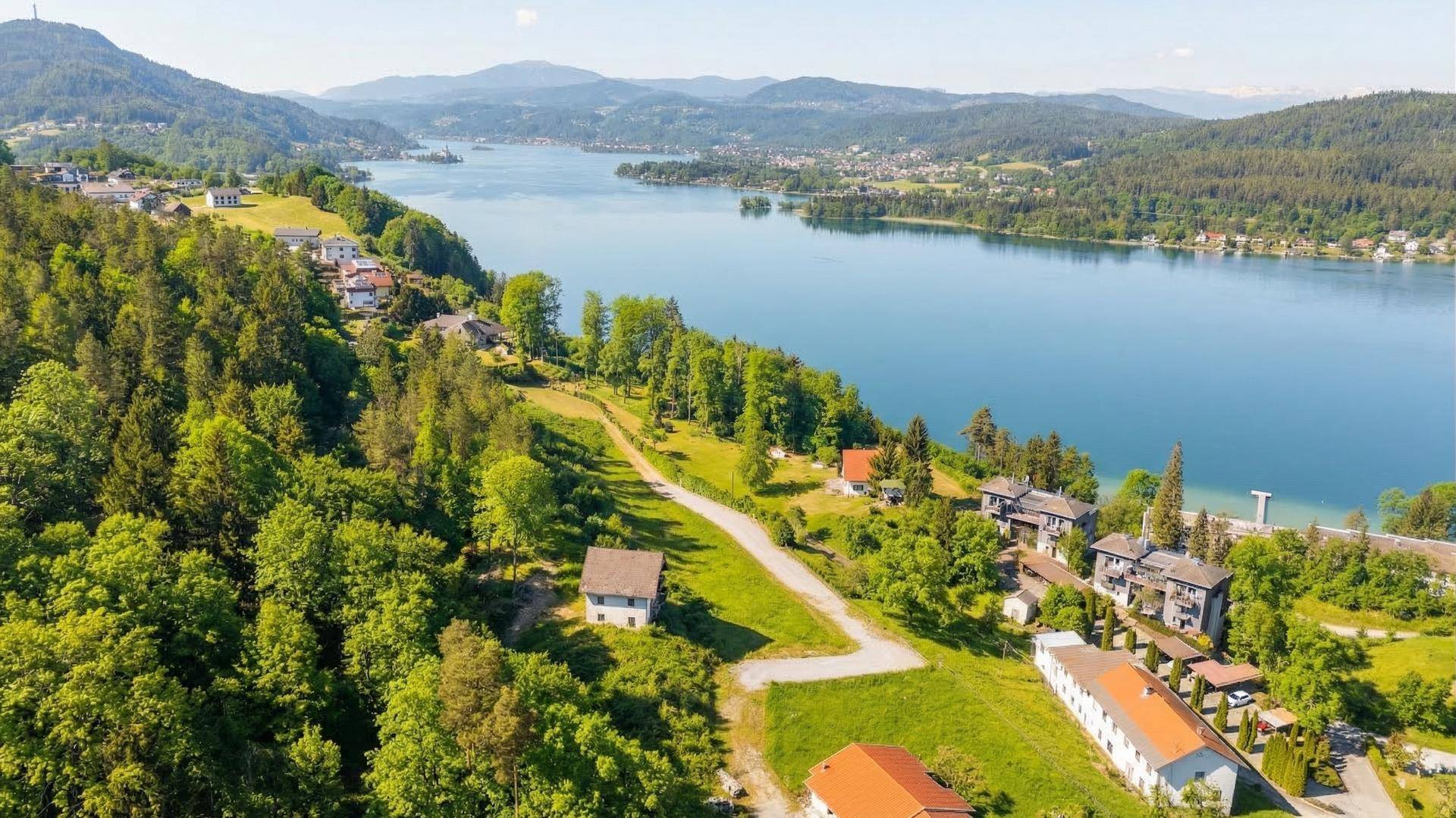 Panoramablick auf eine Siedlung am Seeufer mit bewaldeten Hügeln und Bergen im Hintergrund.