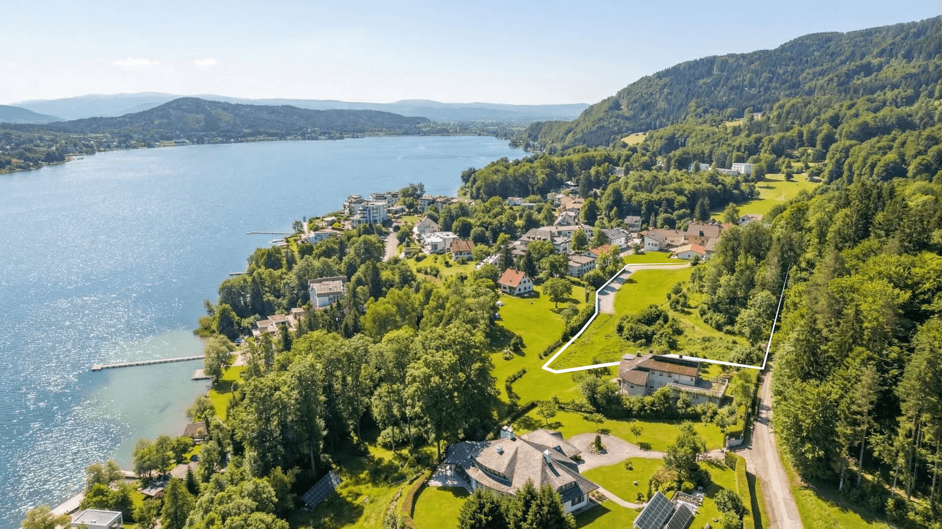 Luftaufnahme einer Siedlung am Seeufer mit umrandetem Grundstück und Panoramablick auf die Umgebung.