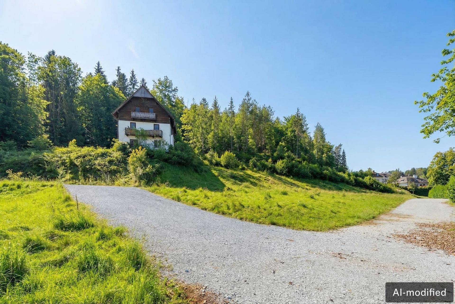 Haus auf einem grünen Hügel mit Schotterweg und bewaldeter Umgebung unter blauem Himmel.
