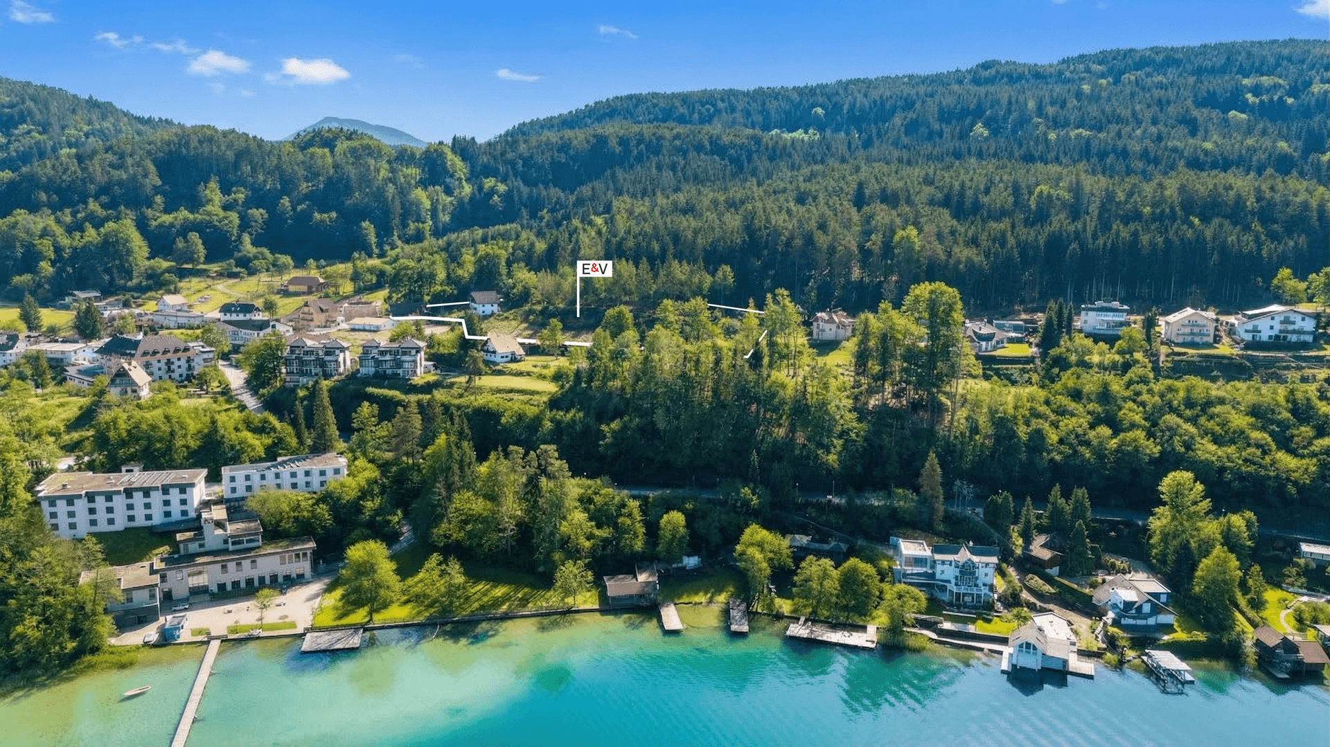 Weitläufige Luftaufnahme des Wörthersees mit Uferbebauung und bewaldeten Hängen unter strahlendem Himmel.