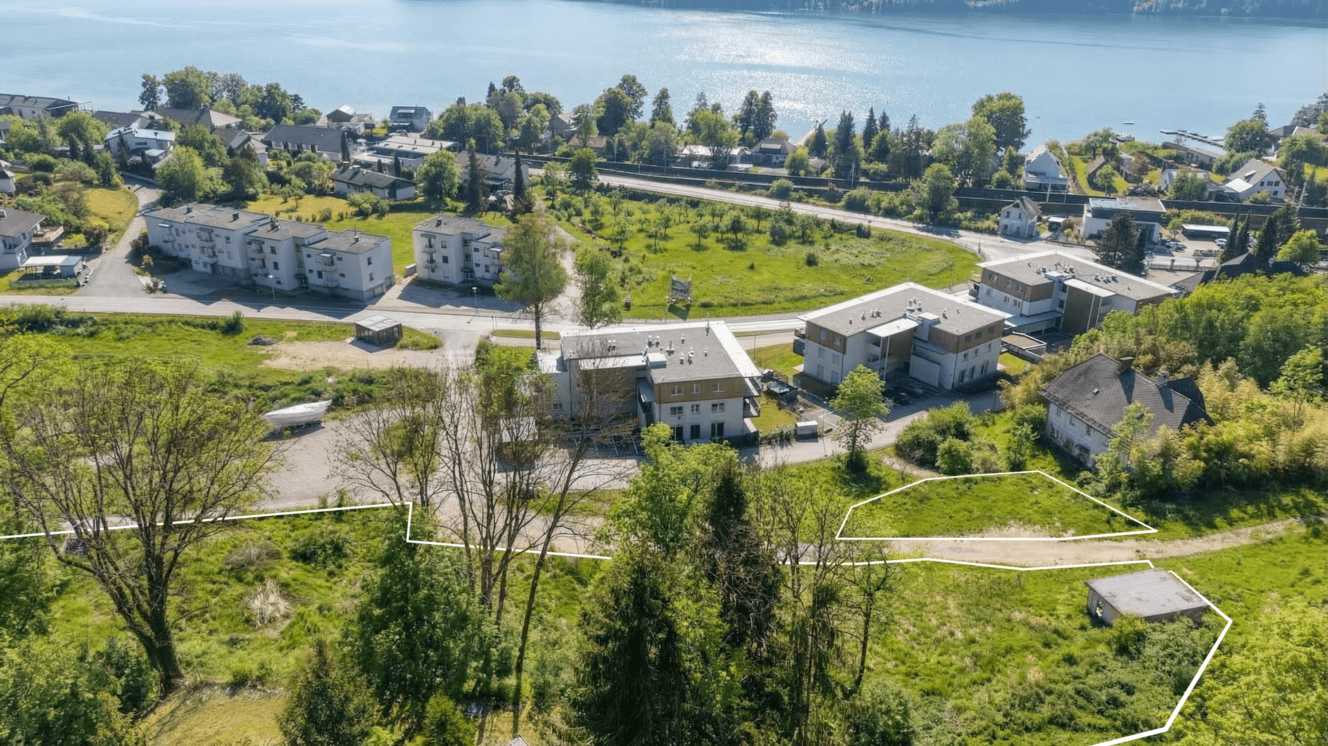 Luftaufnahme eines Grundstücks mit weißer Umrandung, Gebäuden, Grünflächen und Seeblick.