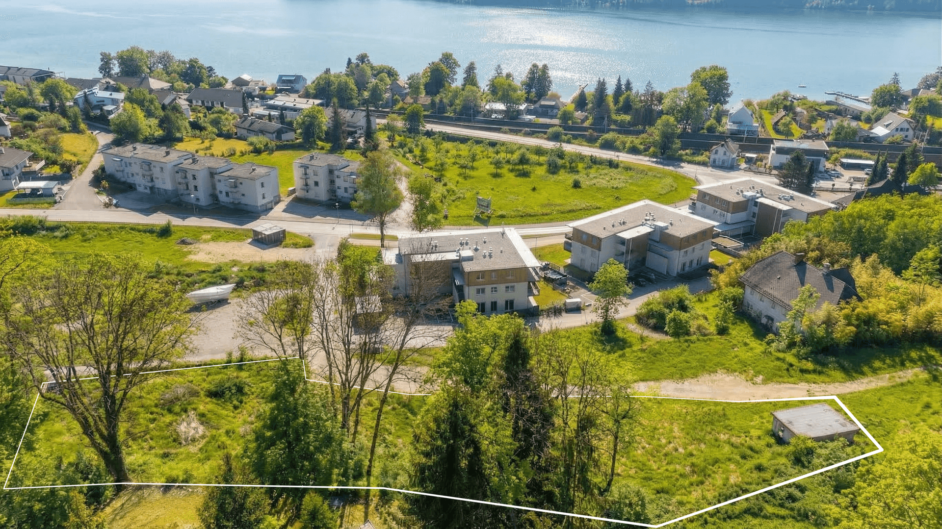 Luftaufnahme eines Grundstücks mit weißer Umrandung, Gebäuden, Grünflächen und Seeblick.