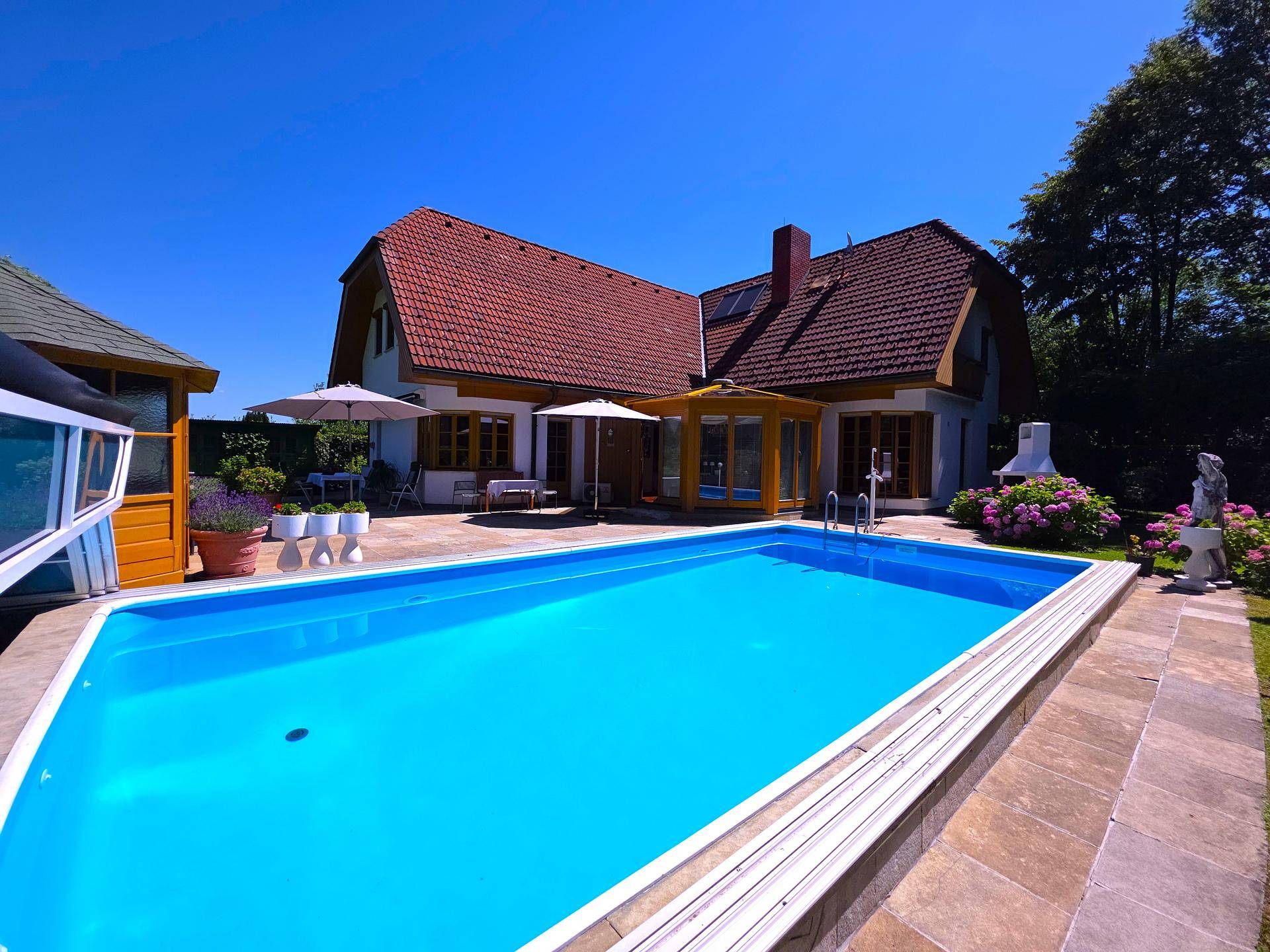 Sonniger Garten mit großem Pool, gepflasterter Terrasse und blühenden Pflanzen.