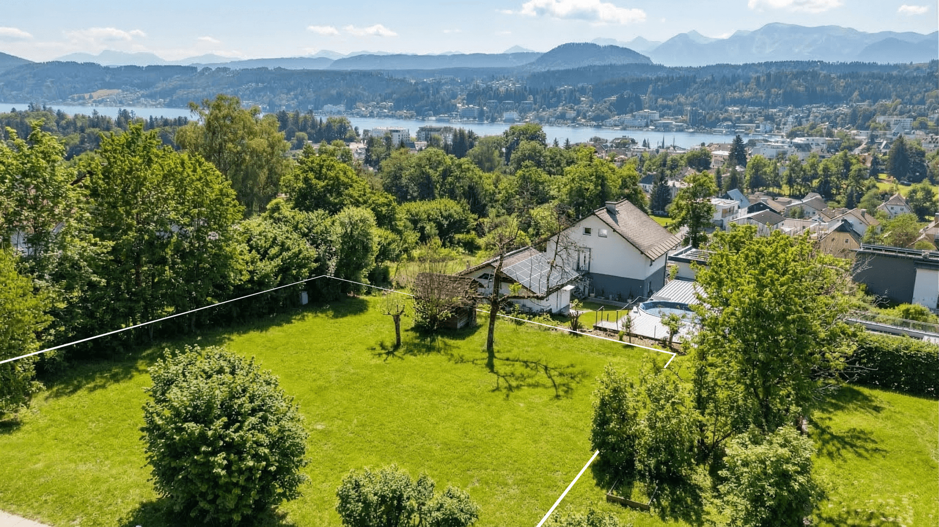 Luftaufnahme eines Anwesens mit großem Rasen, Bäumen, einem See und Bergen im Hintergrund, umgeben von Natur.