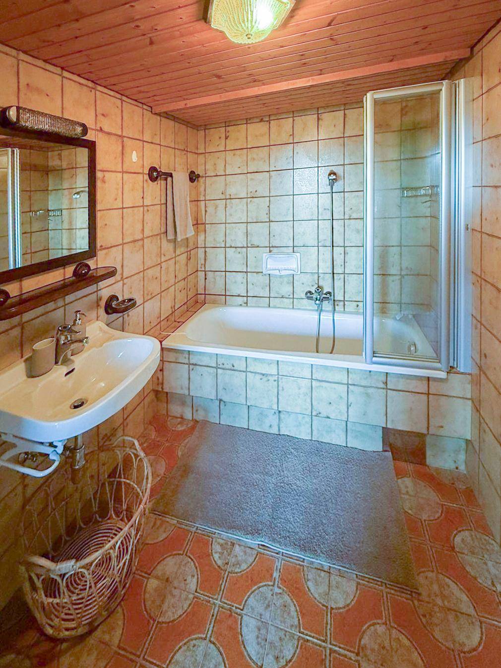 Badezimmer im 80er-Jahre-Stil mit beigen Fliesen, Badewanne mit Duschabtrennung und Einzelwaschbecken.