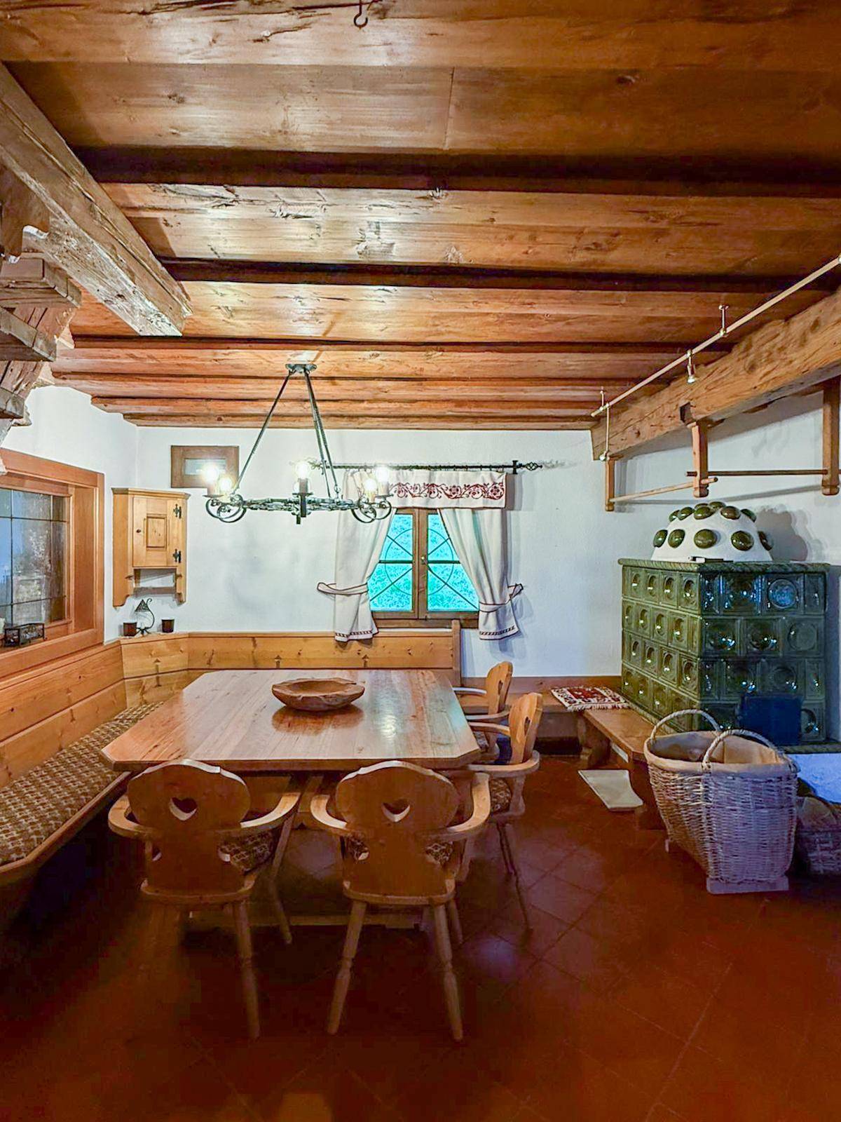 Rustikaler Essbereich mit Holztisch, Eckbank, sichtbaren Deckenbalken und einem grünen Kachelofen.