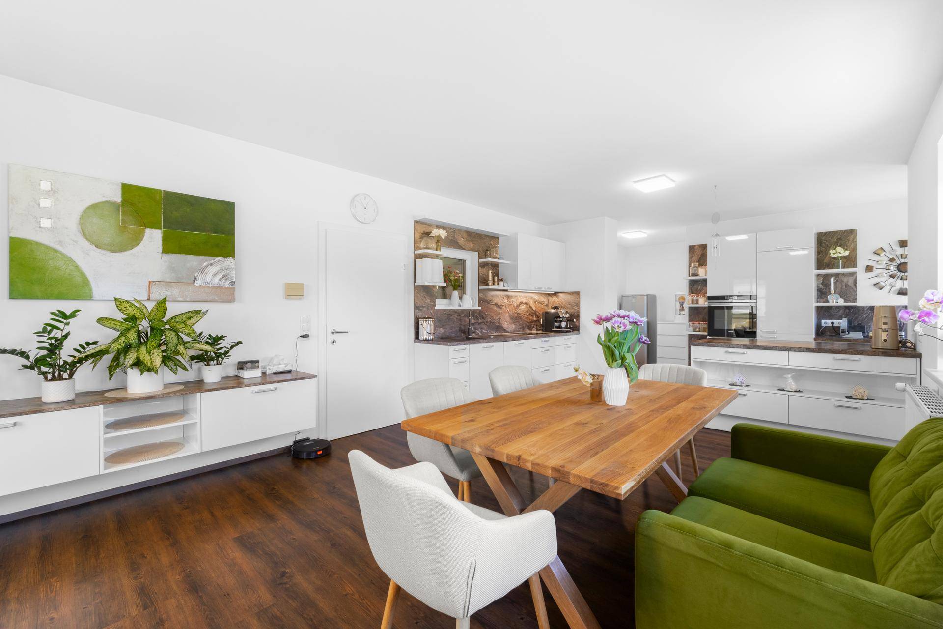 Heller offener Wohn-Essbereich mit grüner Couch, Holztisch und moderner weißer Einbauküche.