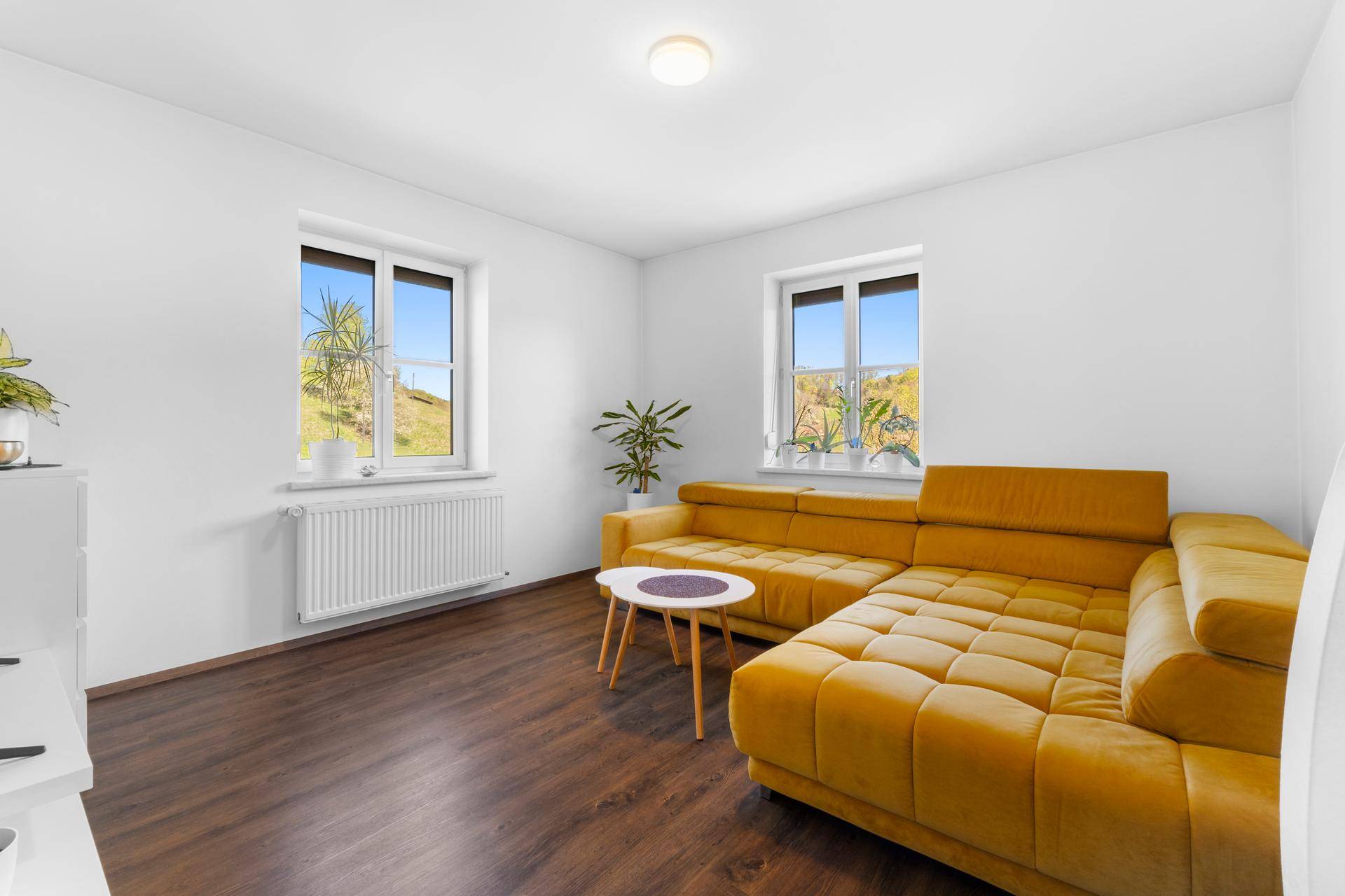 Gemütliches Wohnzimmer mit großem gelbem Ecksofa und zwei Fenstern mit Blick auf die Landschaft.