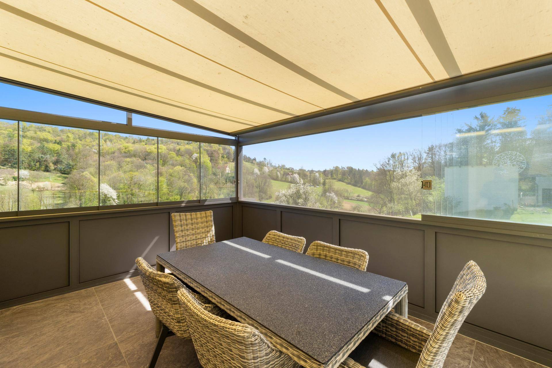 Verglaste Terrasse mit Esstisch und Stühlen, die einen weiten Panoramablick ins Grüne bietet.
