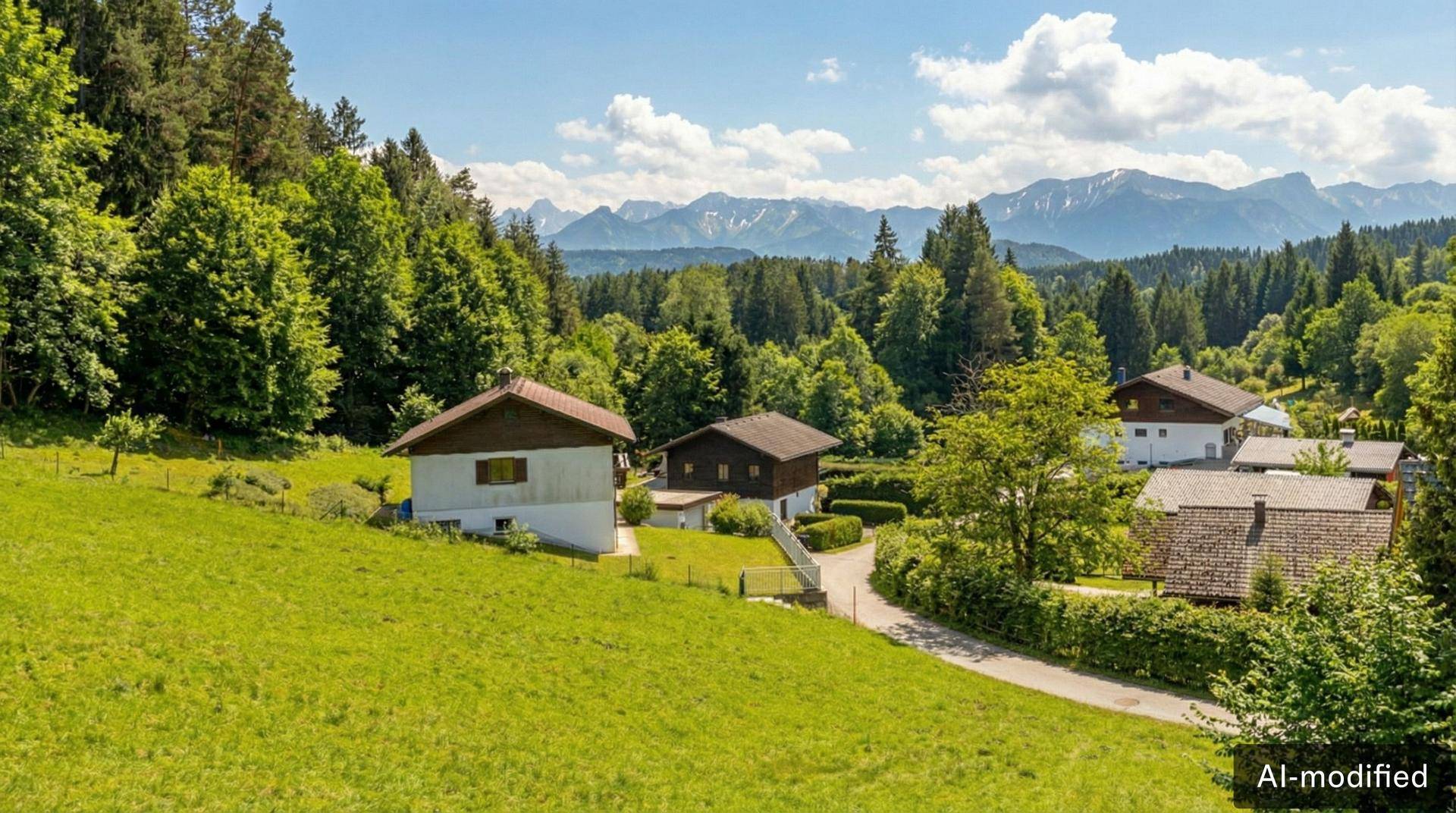 Blick auf Wohnhäuser an einem grünen Hang, umgeben von Wäldern und einer beeindruckenden Bergkulisse.
