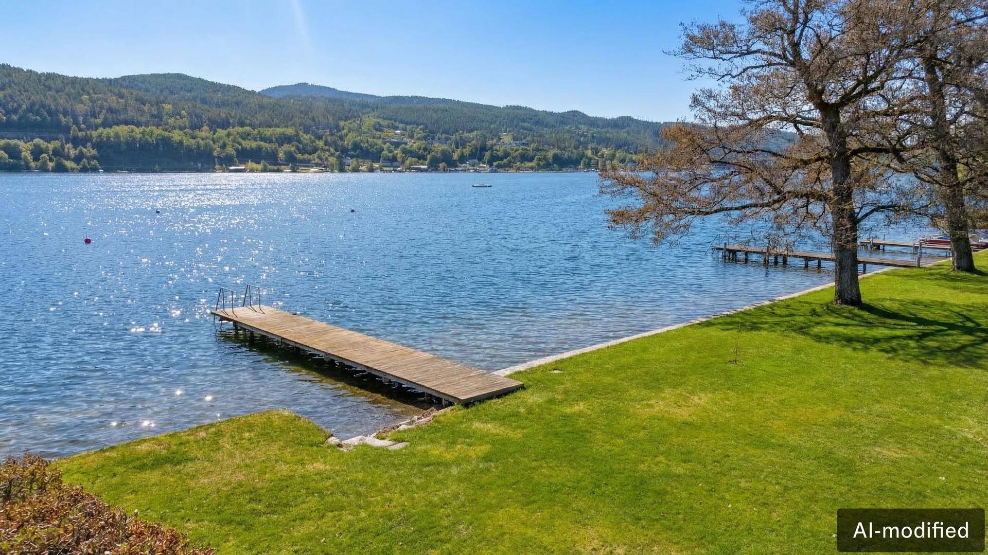 Weitläufige Rasenfläche am Seeufer mit langem Holzsteg und Panoramablick auf die Berge.