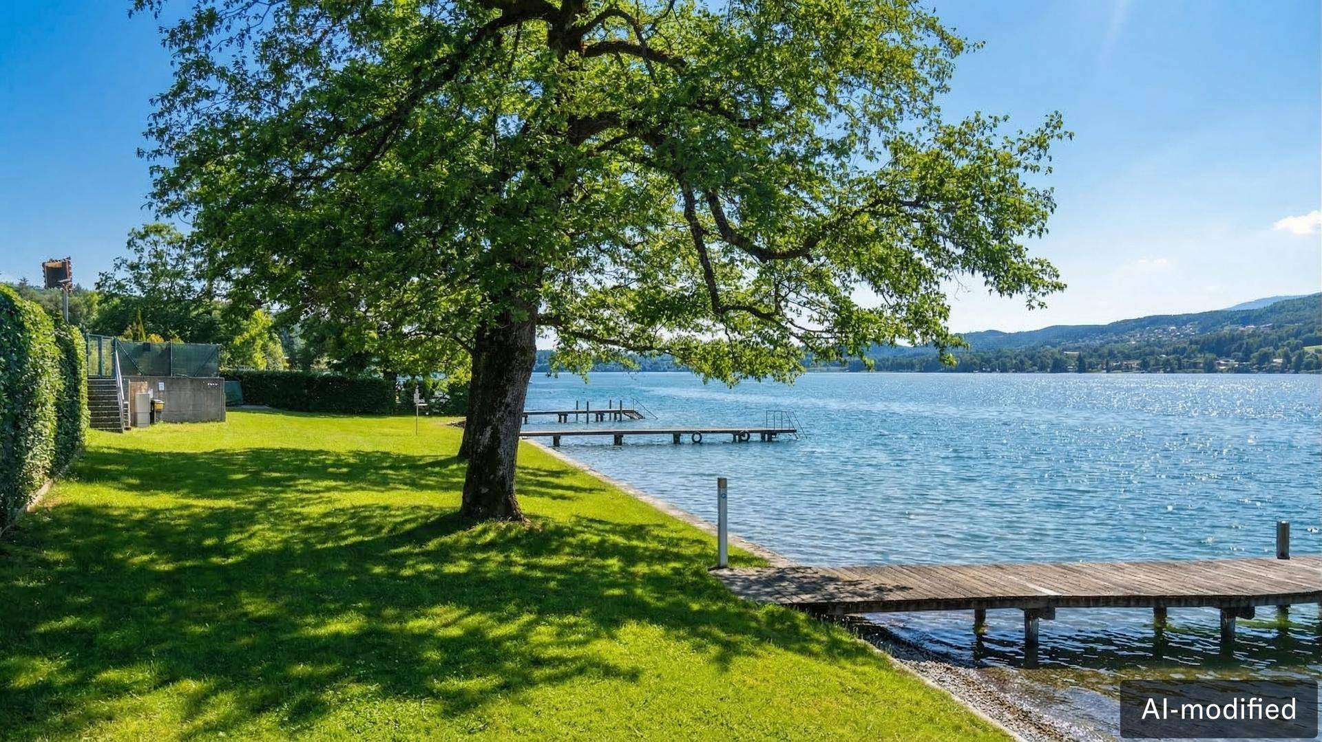 Sonniger Gartenbereich am See mit Holzsteg, großem Baum und Bergblick.