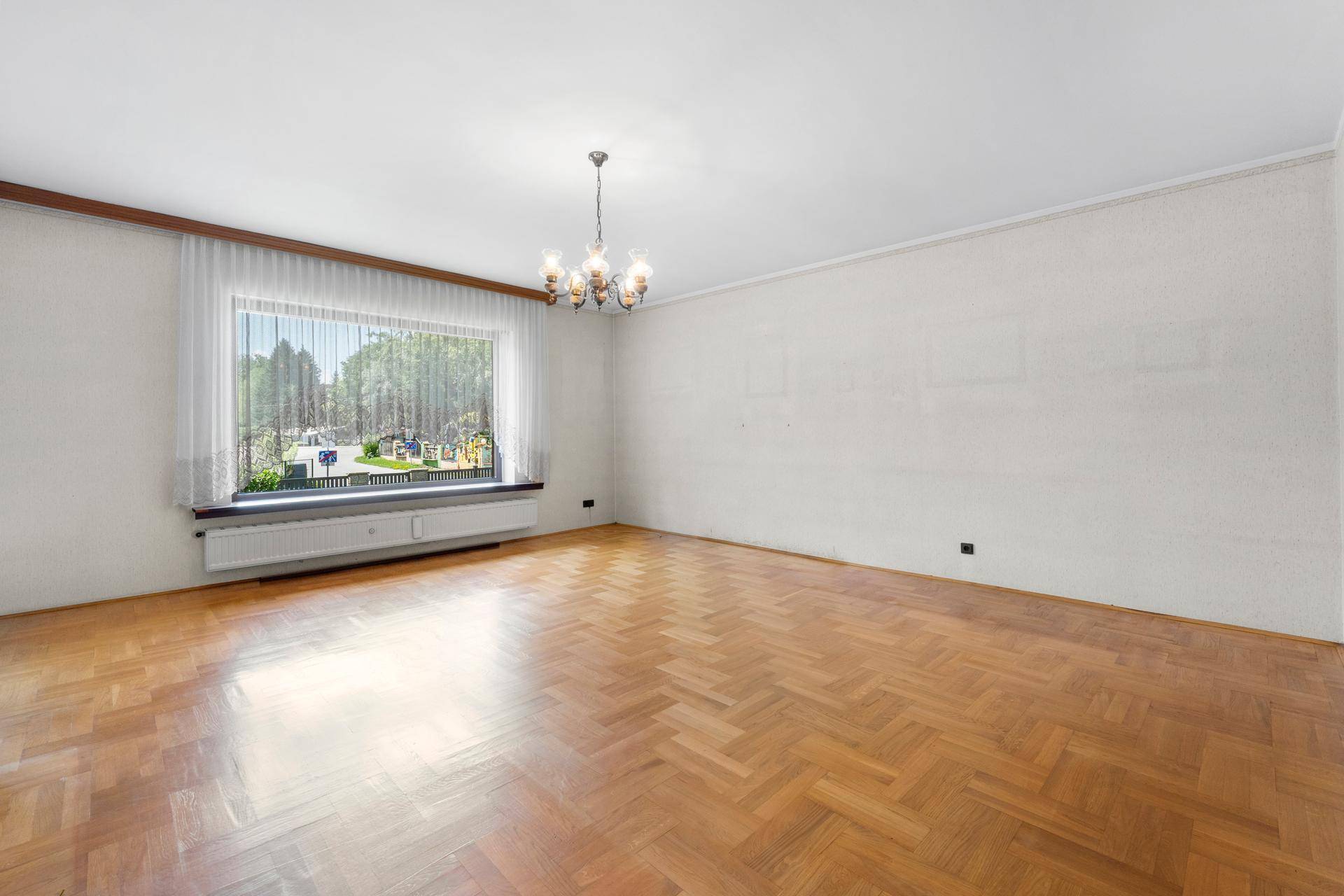 Helles Wohnzimmer mit Fischgrätparkettboden und großem Fenster, das viel Tageslicht hereinlässt.