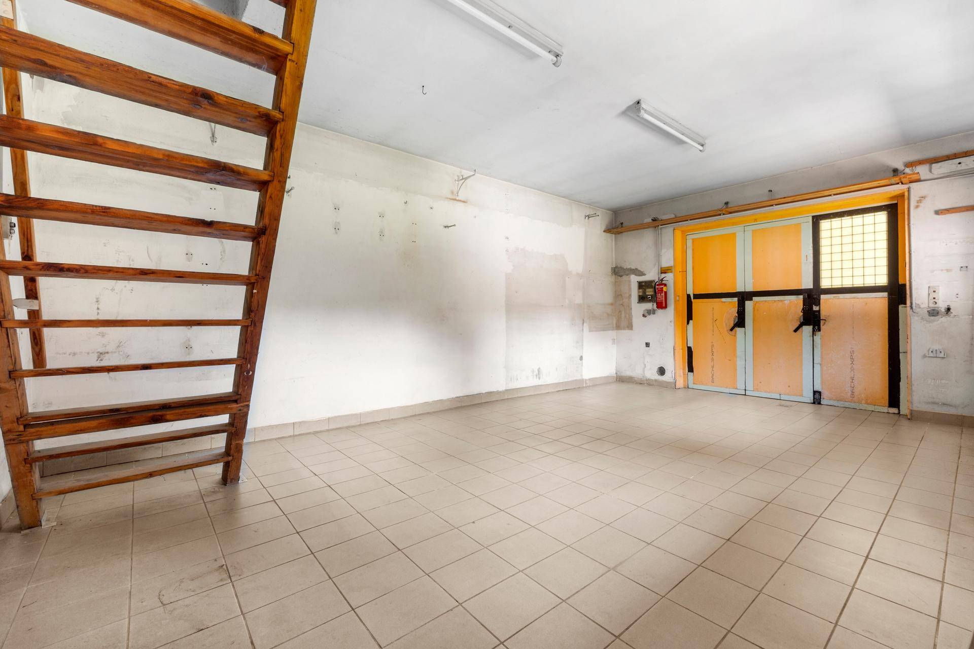 Geräumige Garage mit Fliesenboden, Holztür und einer Holztreppe zu einem oberen Bereich.