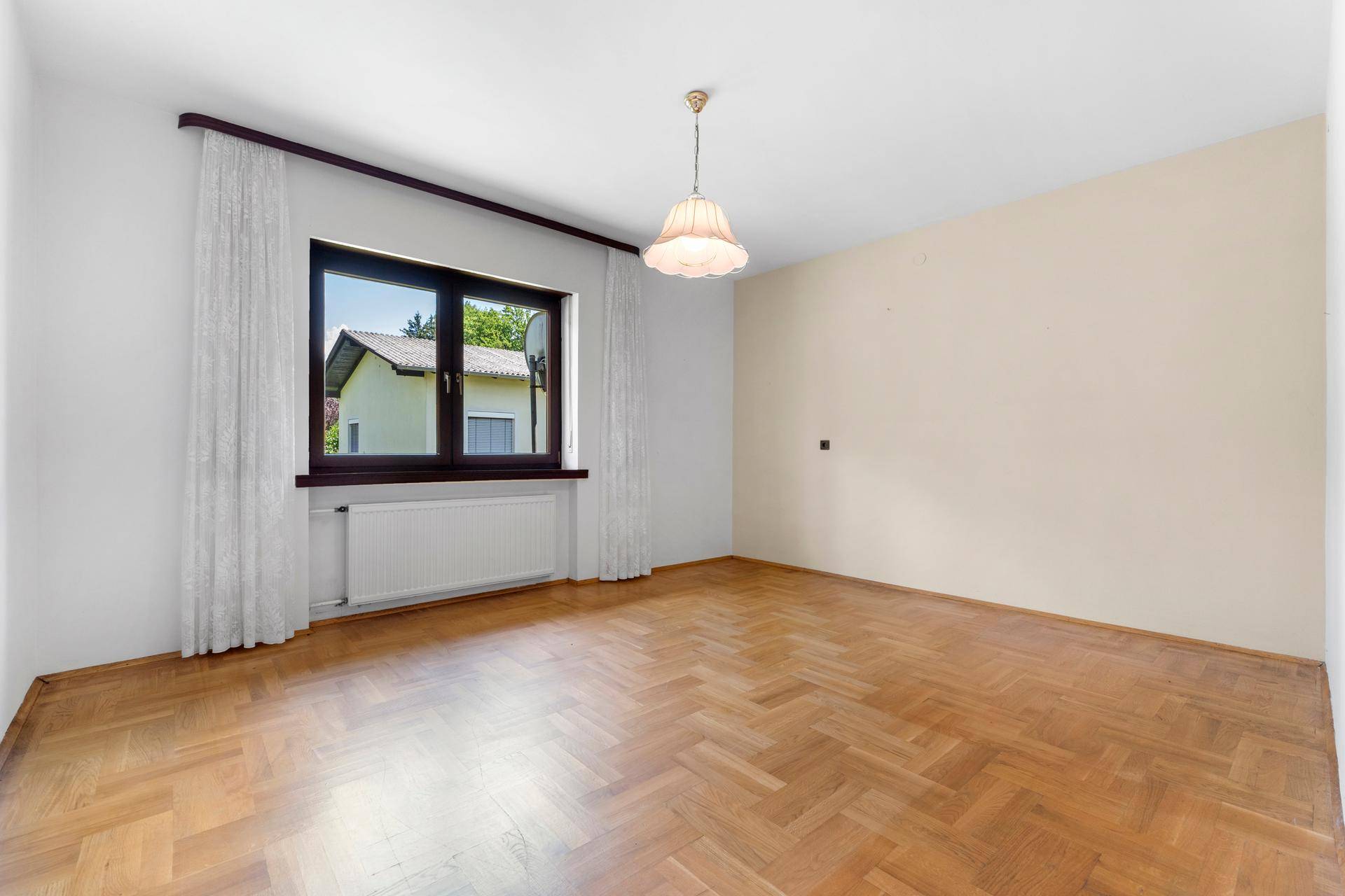 Leeres Zimmer mit Fischgrätparkettboden und einem Fenster, das den Blick ins Grüne freigibt.