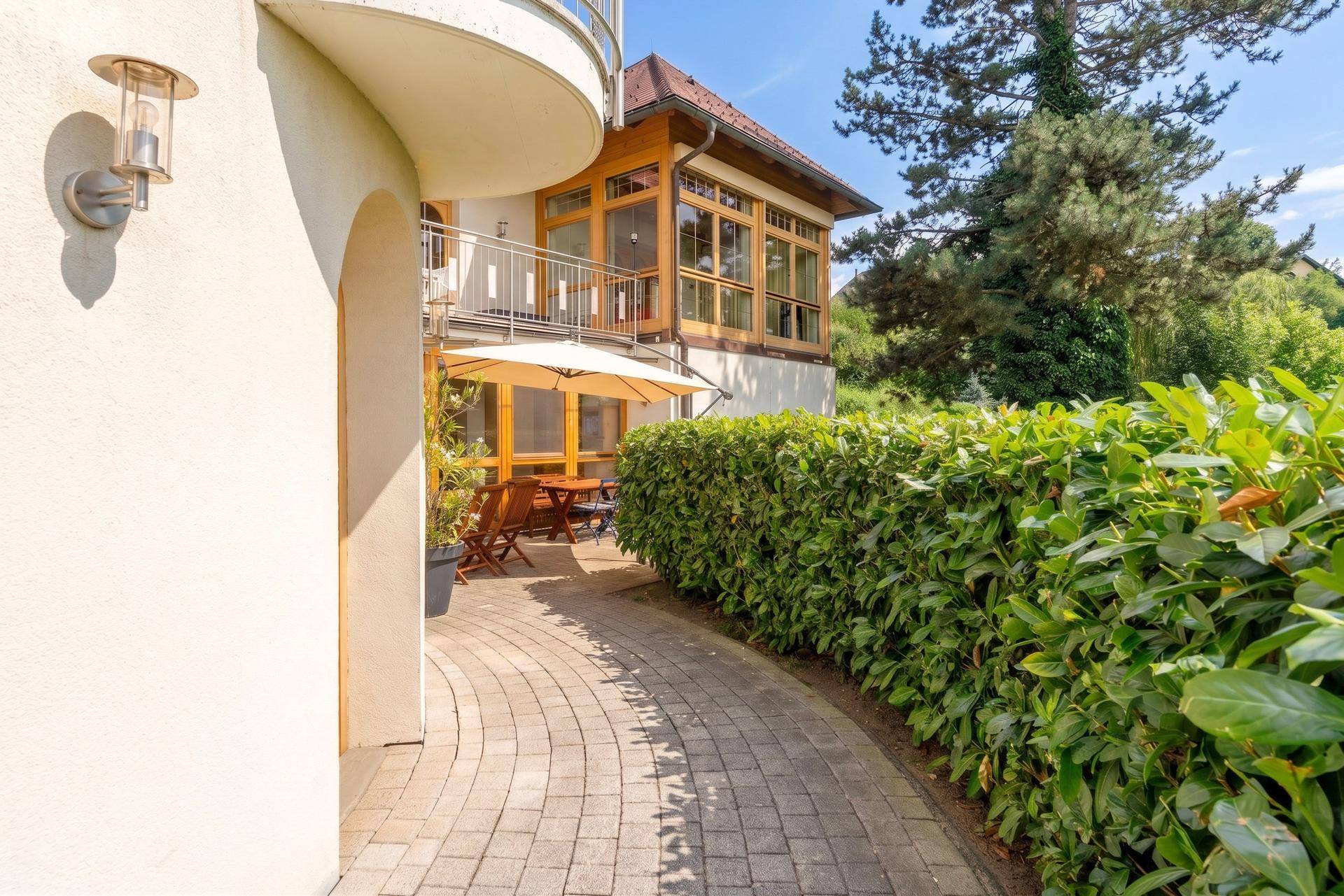 Gepflasterter Weg führt zu einer gemütlichen Terrasse mit Sonnenschirm und Hecke.