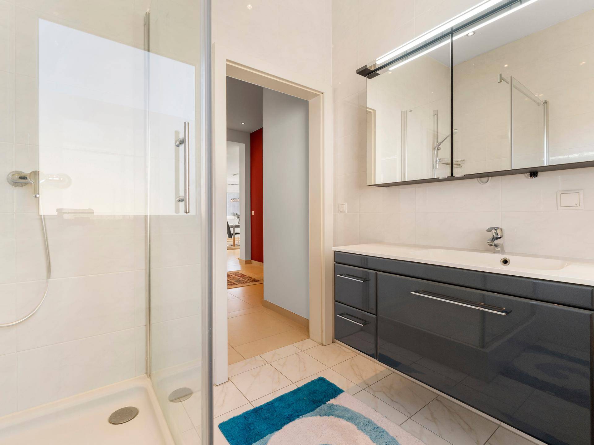 Modernes Badezimmer mit Duschkabine, Waschtischunterschrank und Spiegelschrank, hell gefliest.
