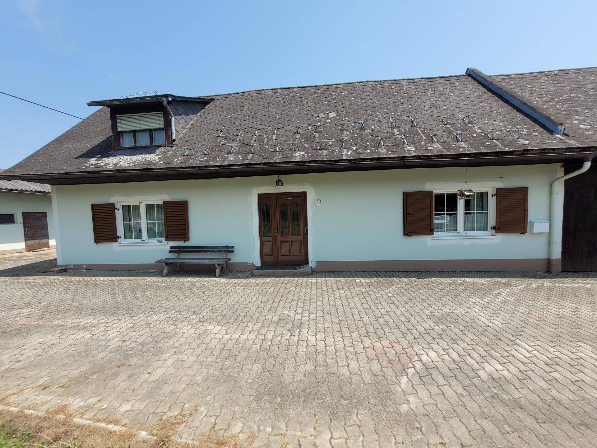 Vorderansicht des Wohnhauses mit hellgrüner Fassade, braunen Fensterläden und gepflastertem Vorplatz.