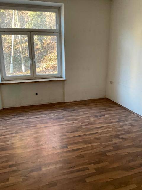 Leeres Zimmer mit Laminatboden und großem Fenster, das viel Licht hereinlässt.