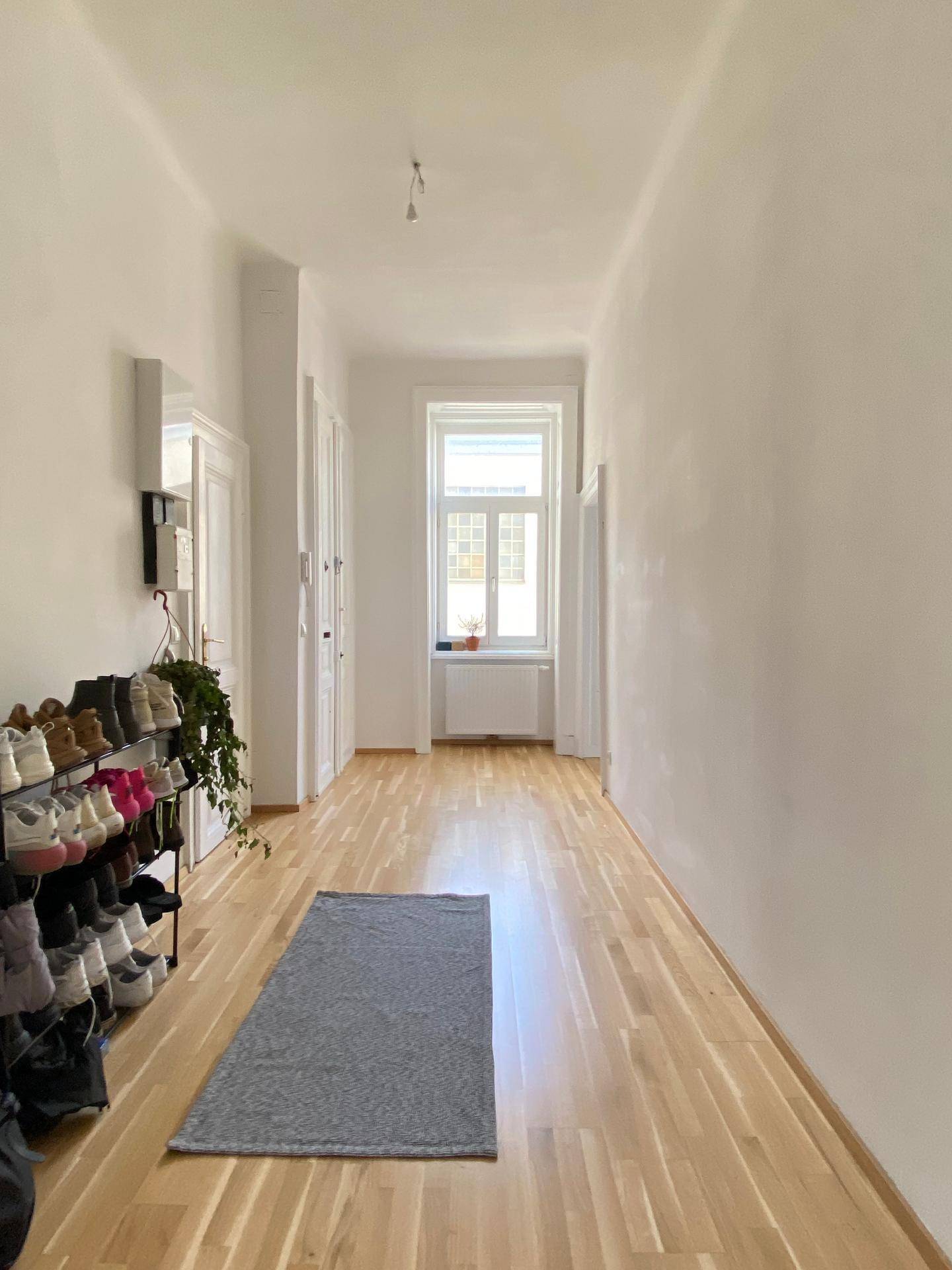 Langer, heller Flur mit Holzboden, Schuhregal und einem Fenster am Ende.