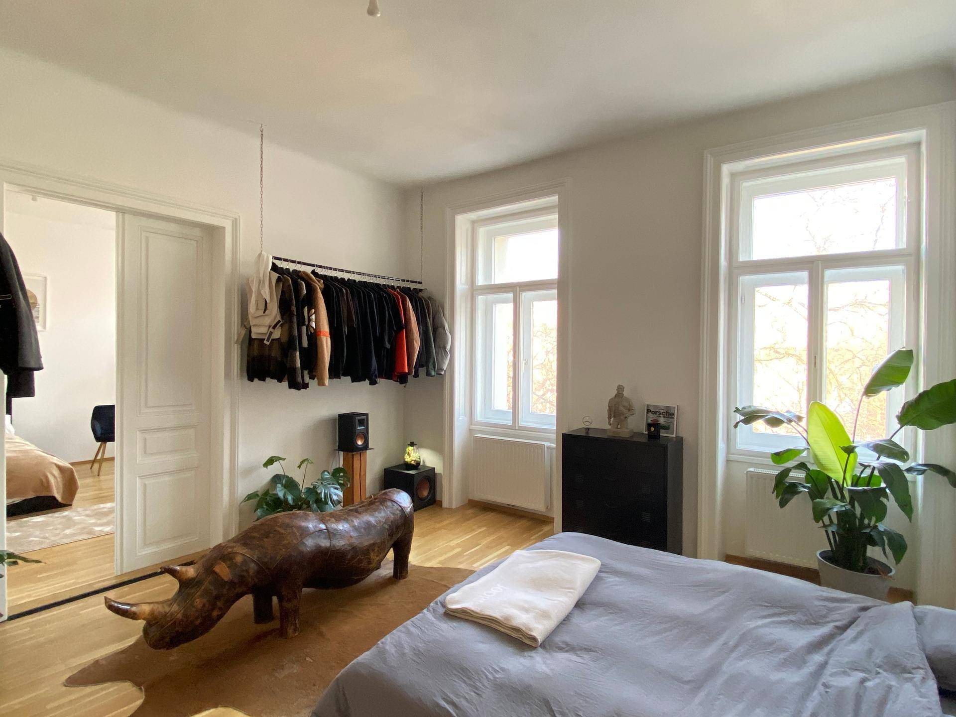 Helles Schlafzimmer mit Kleiderstange, Holzboden und Blick in ein angrenzendes Zimmer.