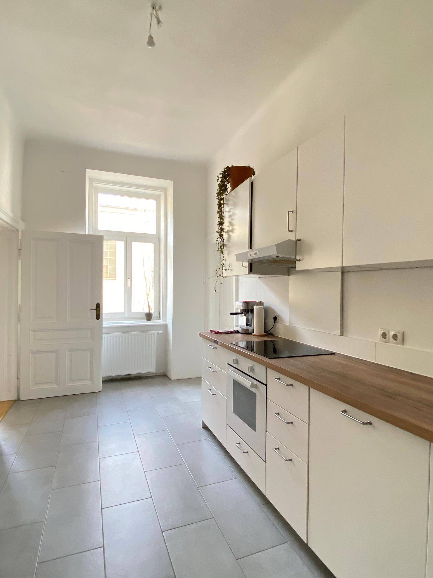 Helle Küche mit weißen Schränken, Holz-Arbeitsplatte, Fliesenboden und Fenster.