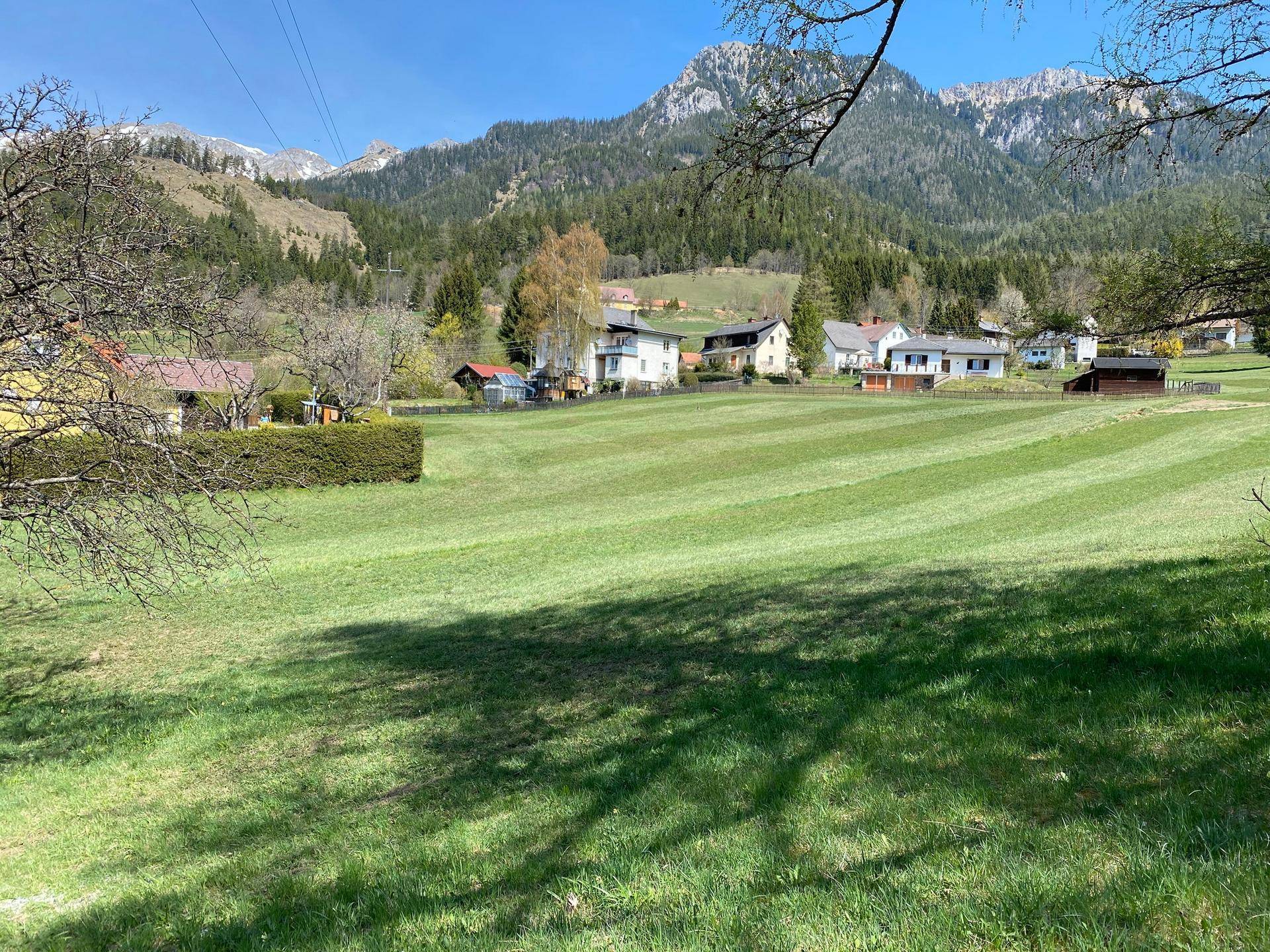 Sonnige Wiesen mit Schattenwurf von Bäumen und Blick auf eine Bergkette und Häuser.