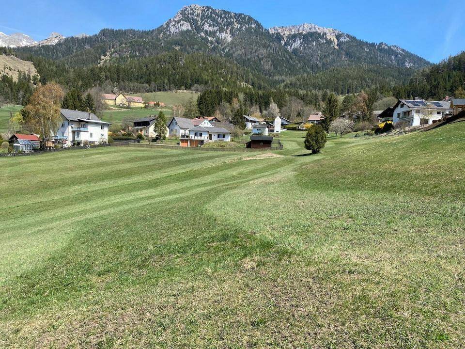 Sonnige Wiesenfläche mit Blick auf markante Felsformationen und eine ländliche Siedlung.