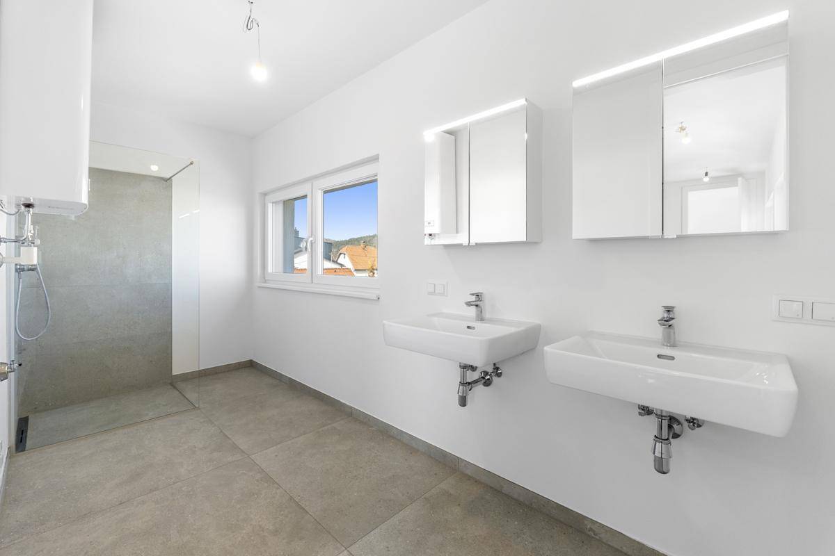 Modernes Badezimmer mit zwei Waschbecken, großer Dusche und Fenster, das viel Tageslicht hereinlässt.