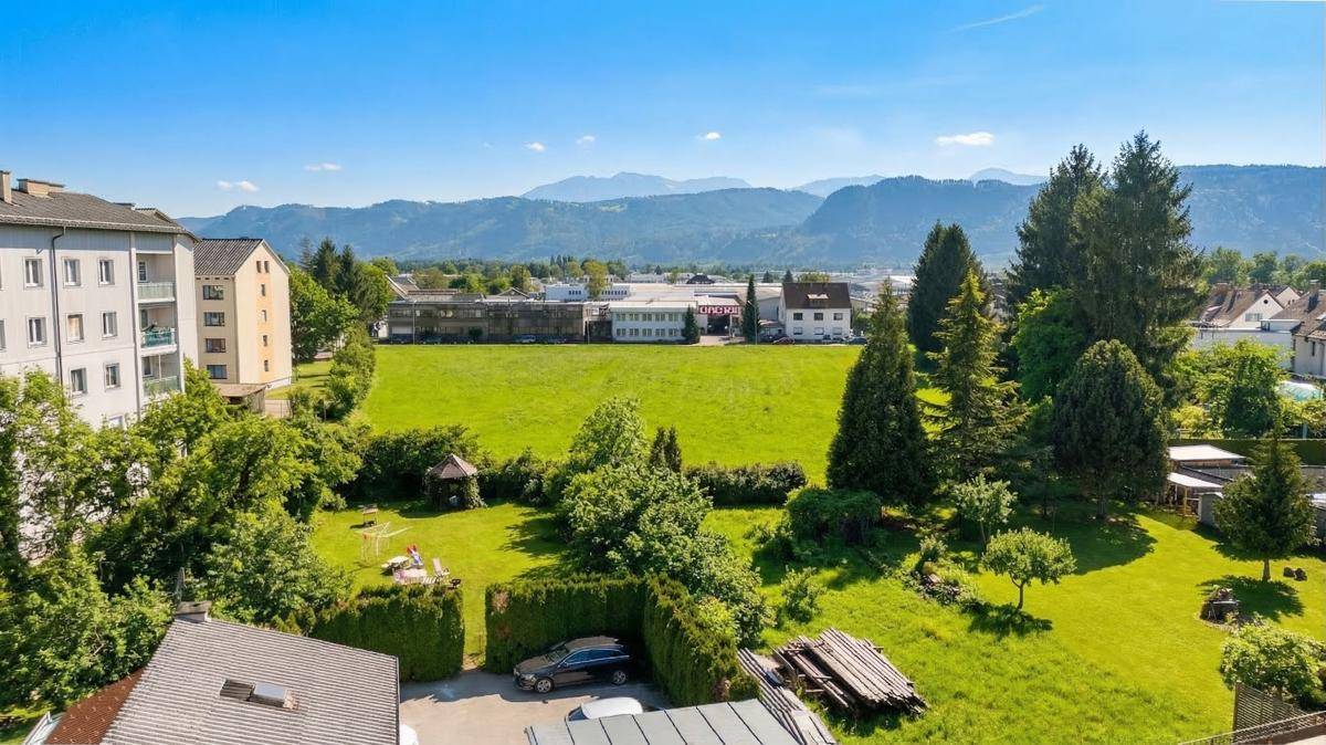 Weitläufige Landschaftsaufnahme mit Grünflächen, Bäumen und Bergen unter klarem blauem Himmel.