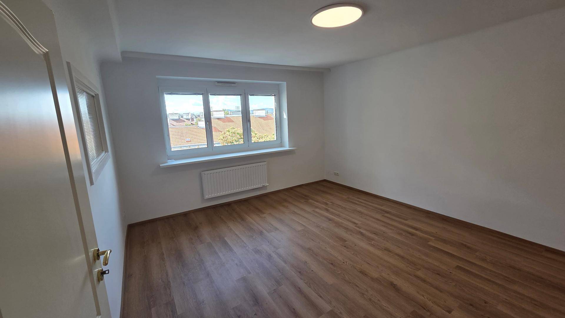 Helles, leeres Zimmer mit Laminatboden, weiß gestrichenen Wänden und einem Fenster mit Stadtblick.