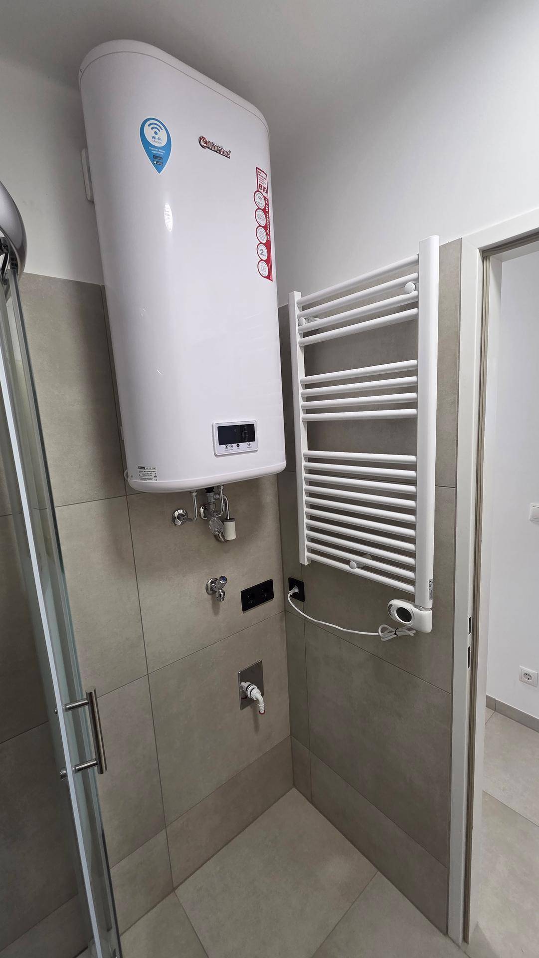 Nahaufnahme eines Heizkörpers und eines Warmwasserboilers in einem gefliesten Badezimmerbereich.