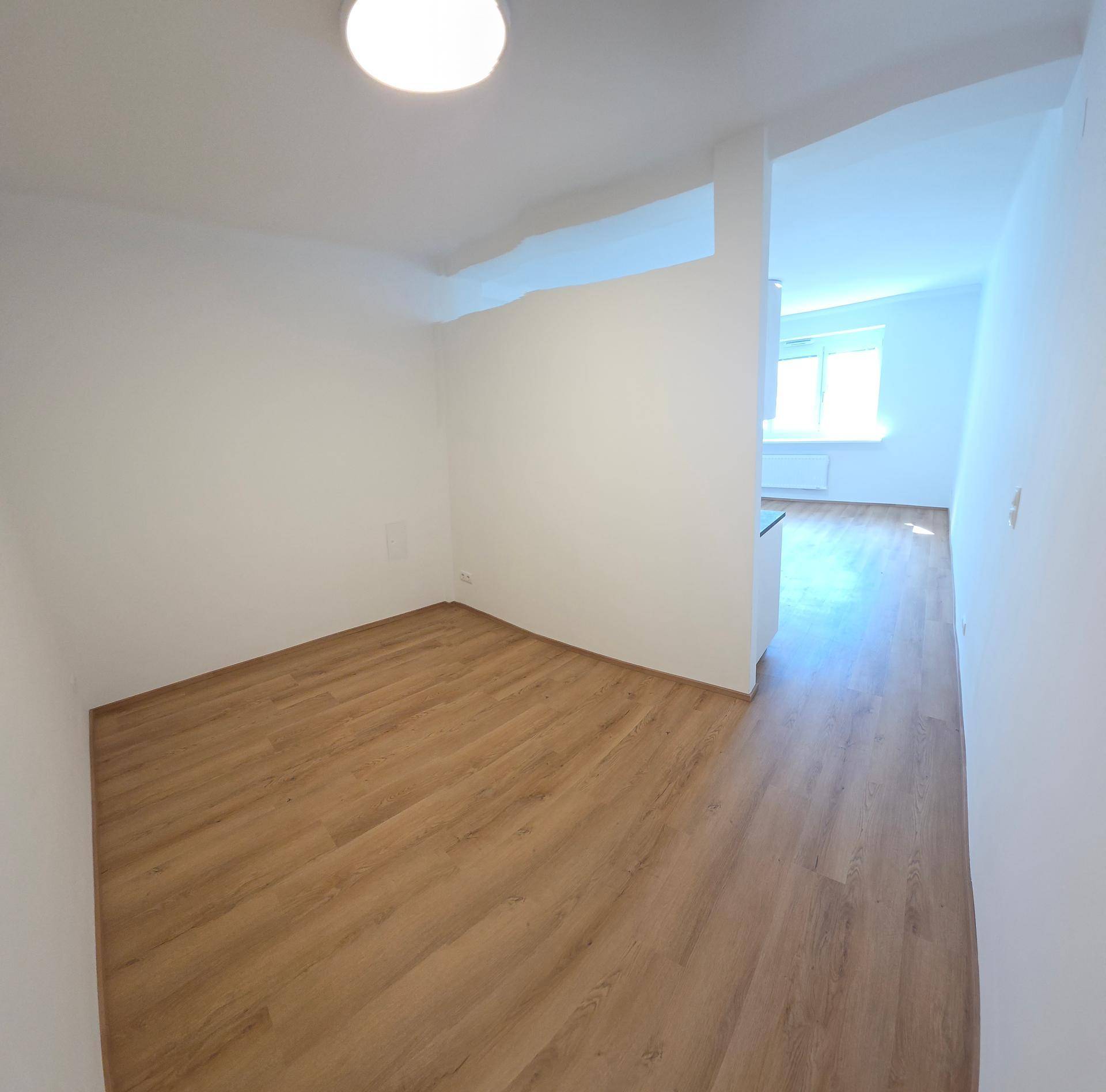 Heller Raum mit Laminatboden und Blick in einen weiteren Bereich mit Fenster und Tageslicht.