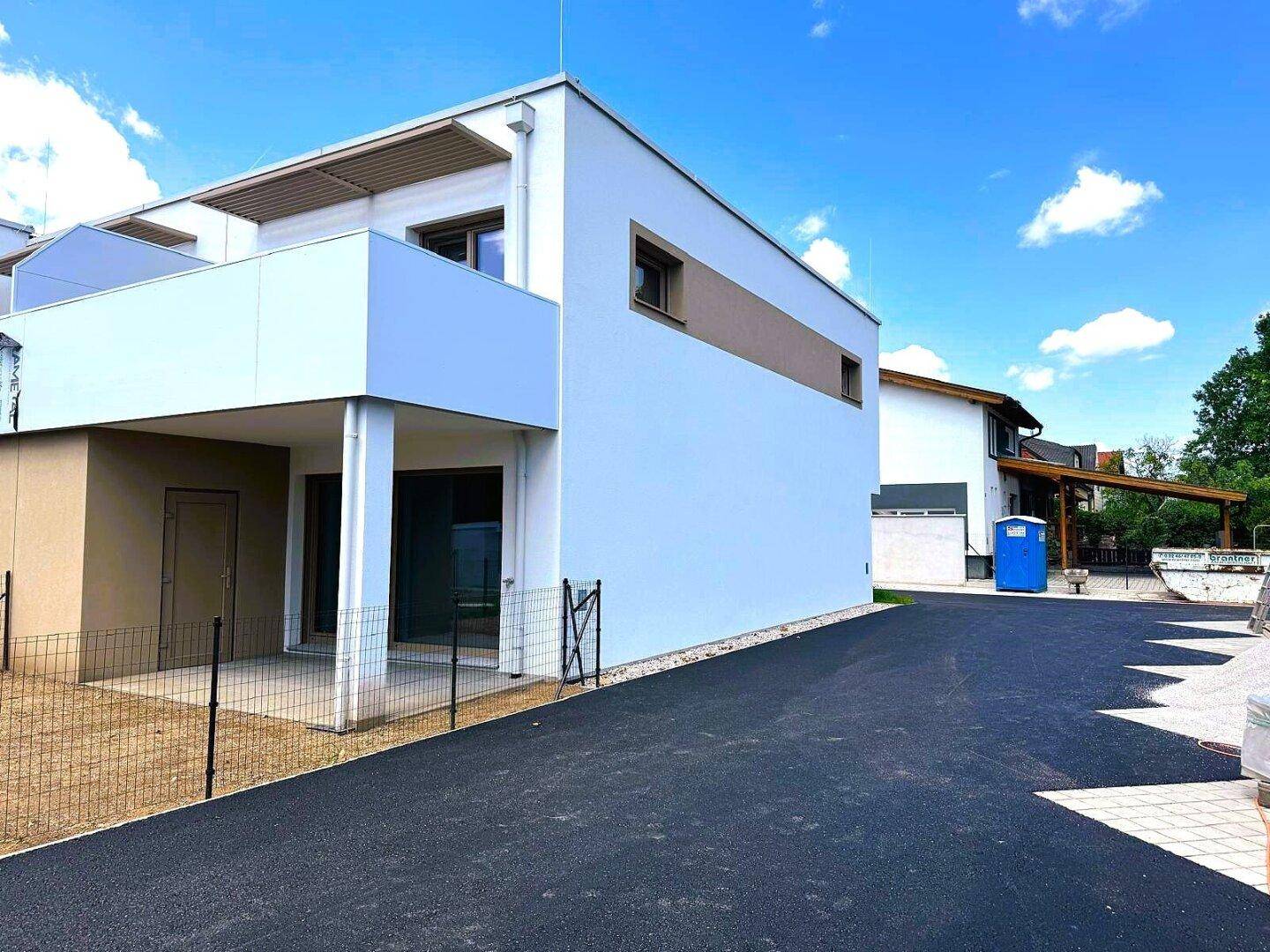 Seitenansicht eines modernen Neubaus mit weißer Fassade und Flachdach, umgeben von einer neuen Zufahrt.
