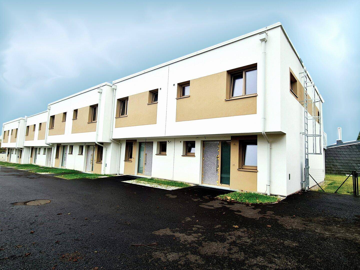 Reihe moderner Häuser mit weißer und beiger Fassade und asphaltierter Zufahrt an einem bewölkten Tag.