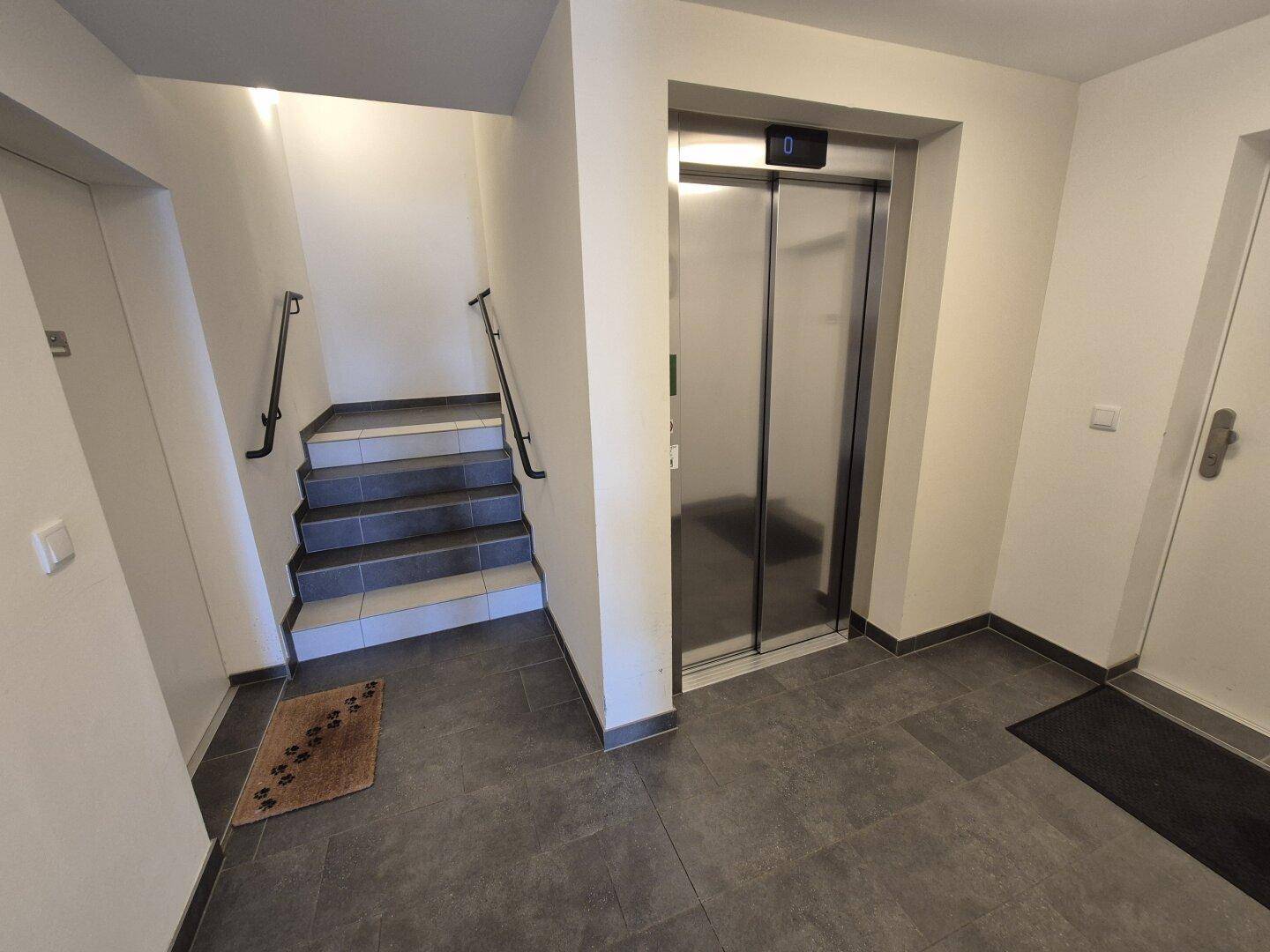 Heller Flur mit modernem Aufzug und Treppenhaus, ausgestattet mit dunklen Bodenfliesen und Handläufen.