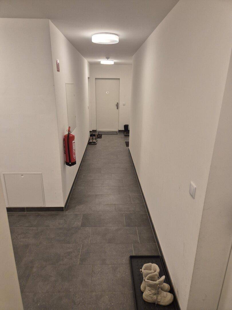 Schmaler Flur mit dunklen Bodenfliesen, weißen Wänden und einem Feuerlöscher an der Wand.