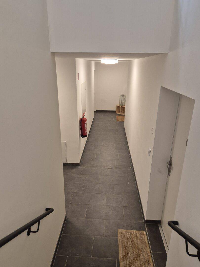 Langer, heller Flur von oben gesehen, mit dunklen Bodenfliesen und Handläufen im Vordergrund.