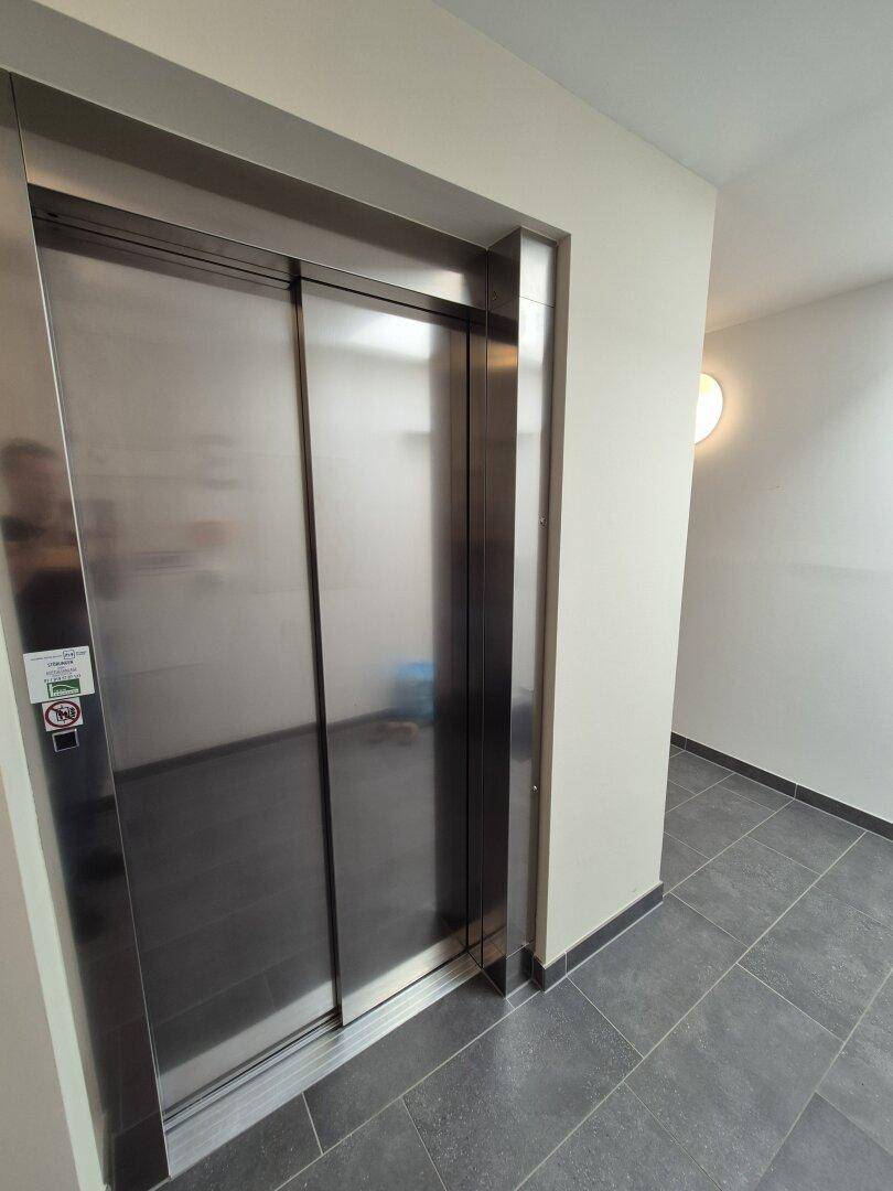Moderner Aufzug mit Edelstahltüren in einem hellen Flur mit dunklen Bodenfliesen.