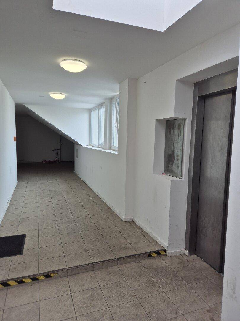 Flur mit Fliesenboden, Aufzug und einer Rampe, die zu einem erhöhten Bereich mit Fenstern führt.