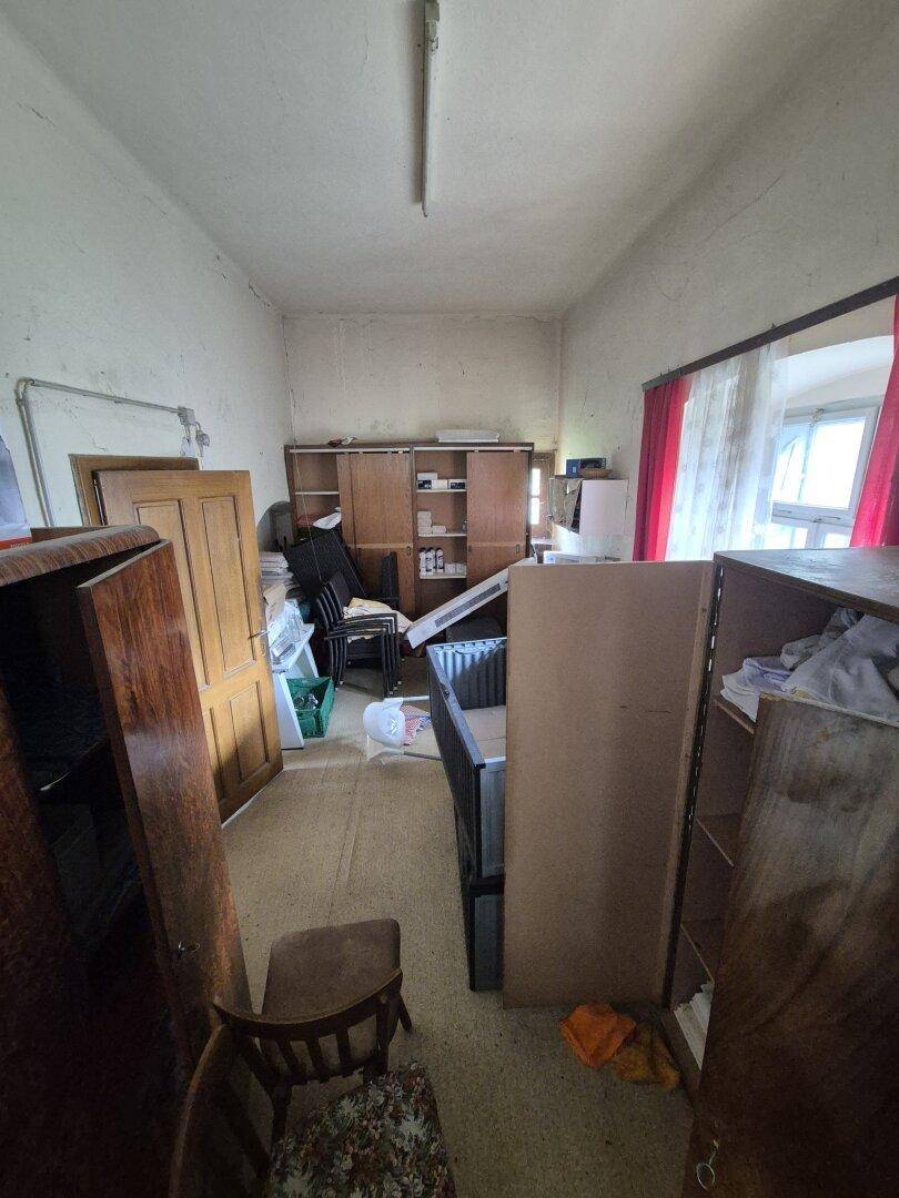 Unaufgeräumter Raum mit Regalen, Schränken und einem Fenster mit roten Vorhängen.