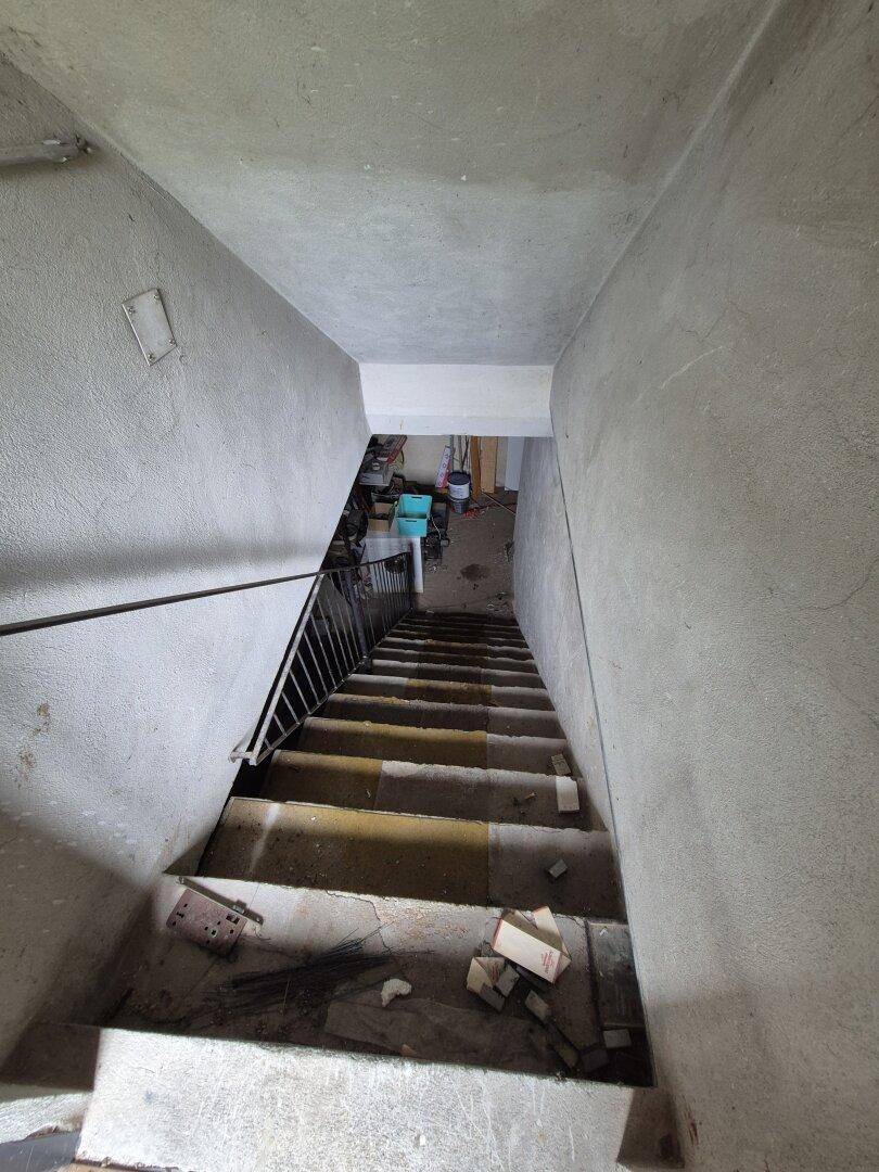 Unfertiges Treppenhaus führt in den Keller, mit Betonwänden und Schutt.