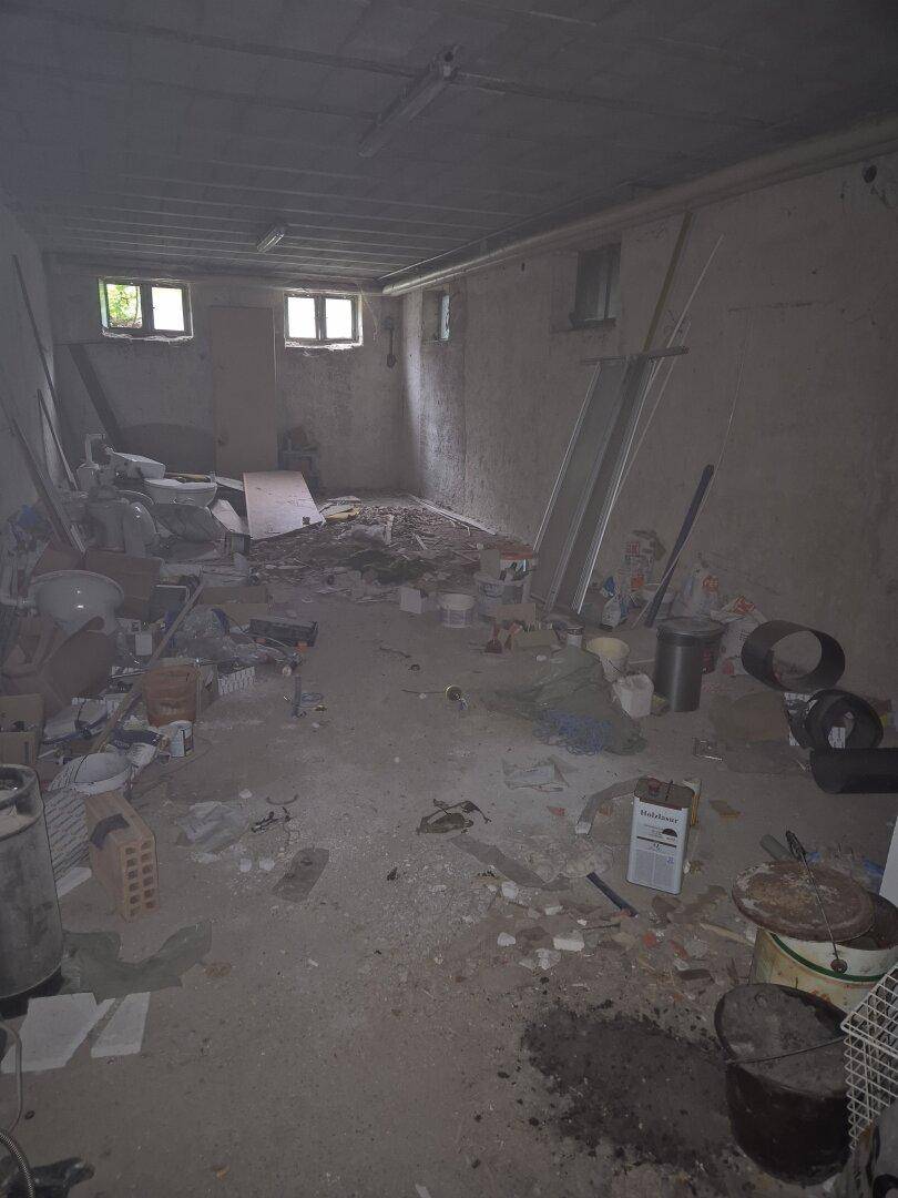 Unaufgeräumter Kellerraum mit Betonwänden, kleinen Fenstern und viel Gerümpel auf dem Boden.
