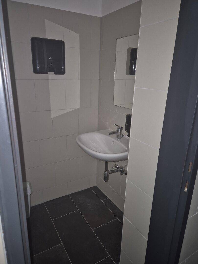 Kleines WC mit Waschbecken, Spiegel und hellgrauen Fliesen an Wänden und Boden.