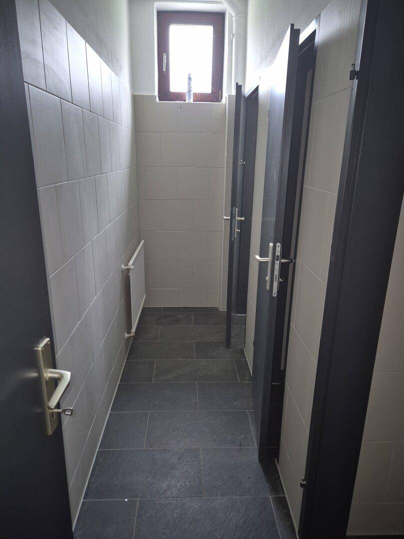 Flur mit mehreren Toilettenkabinen, Fliesenboden und einem kleinen Fenster.