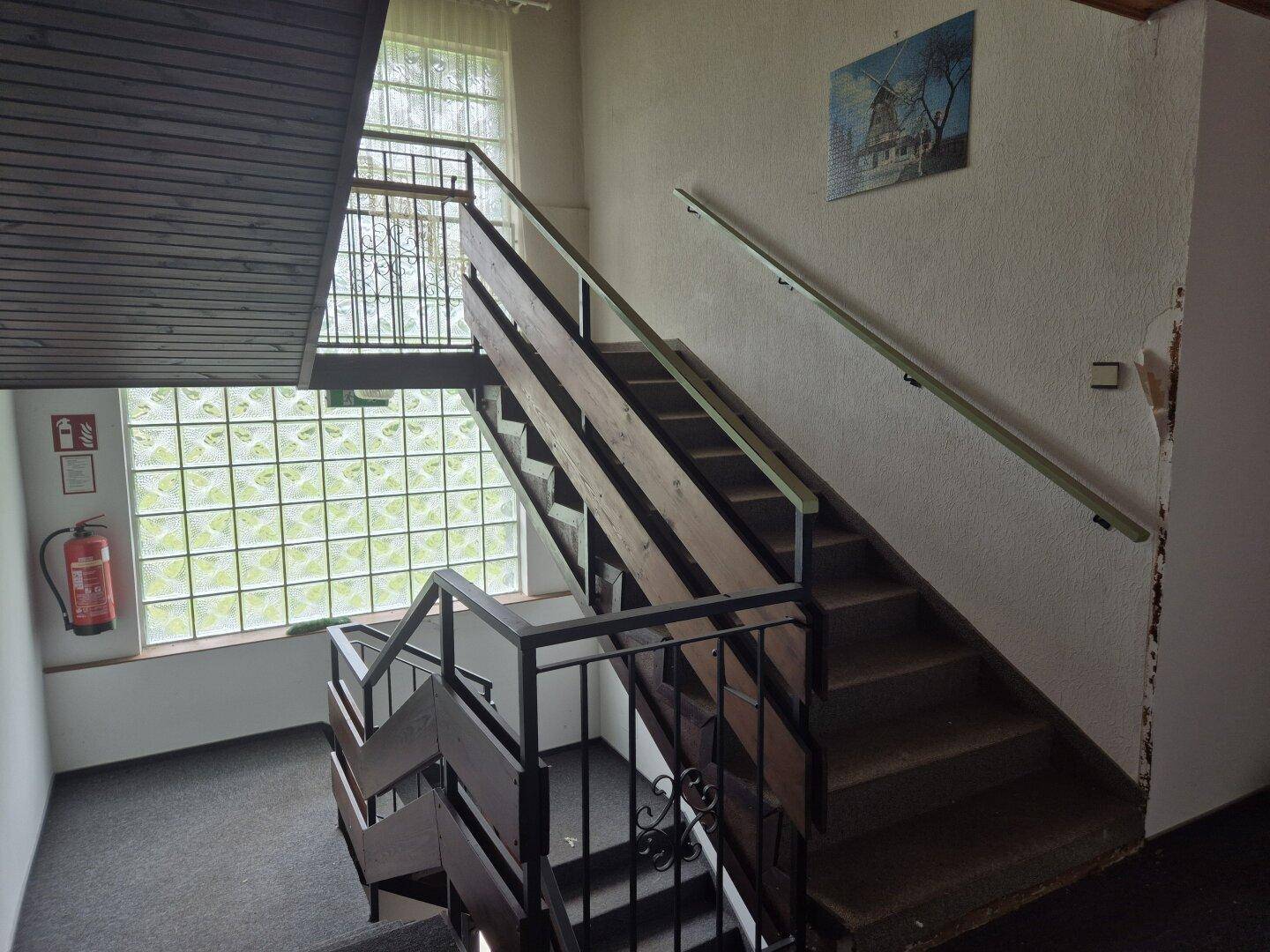 Treppenhaus mit Holzgeländer, Glasbausteinfenstern und Teppichboden auf den Stufen.