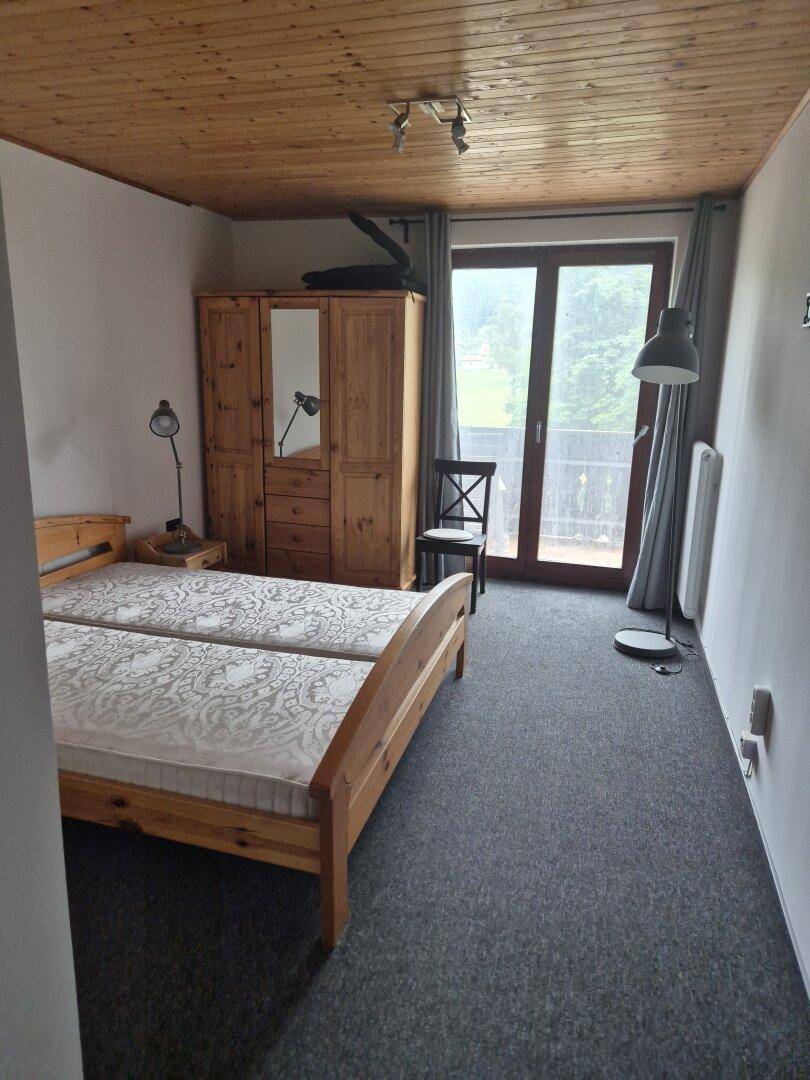 Schlafzimmer mit zwei Einzelbetten, Holzschrank und Balkontür, mit Holzdecke.