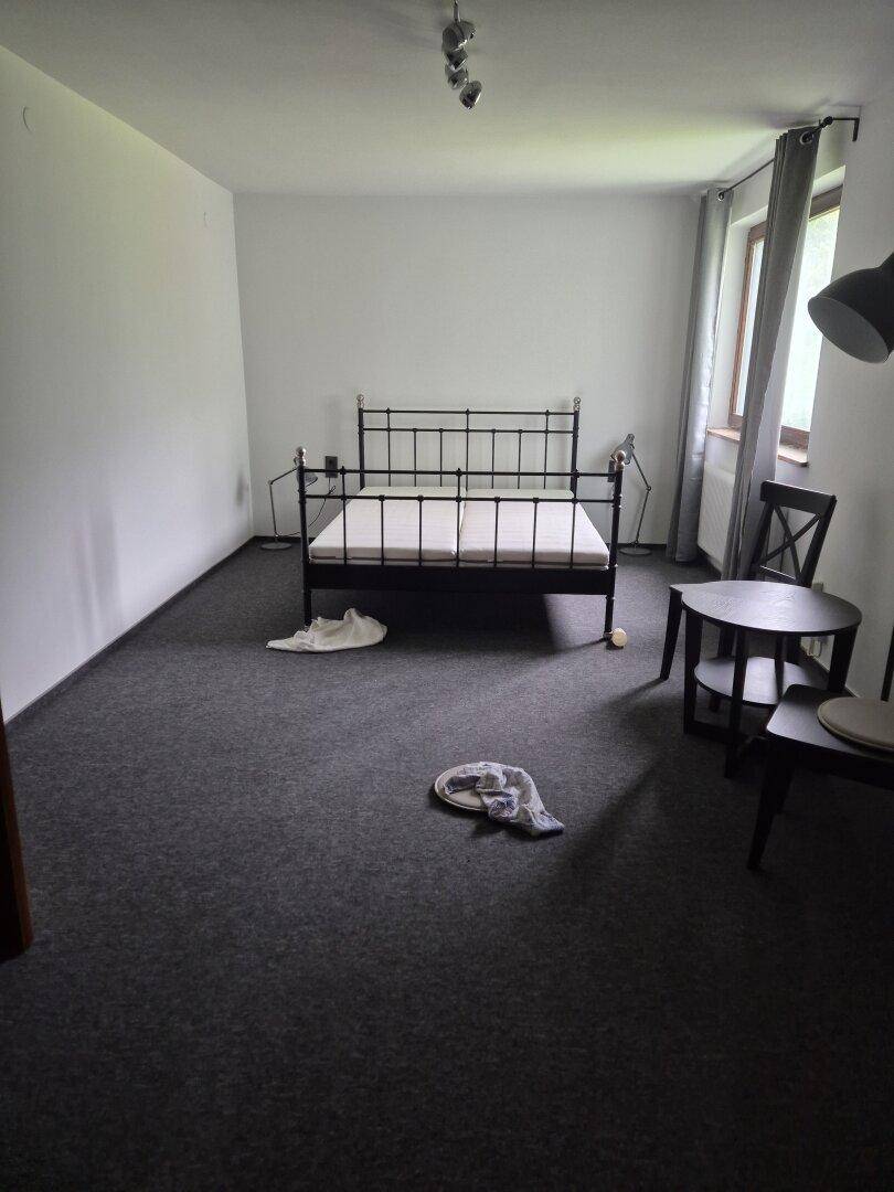 Schlafzimmer mit schwarzem Metallbettgestell, kleinem Tisch und Stühlen auf grauem Teppichboden.