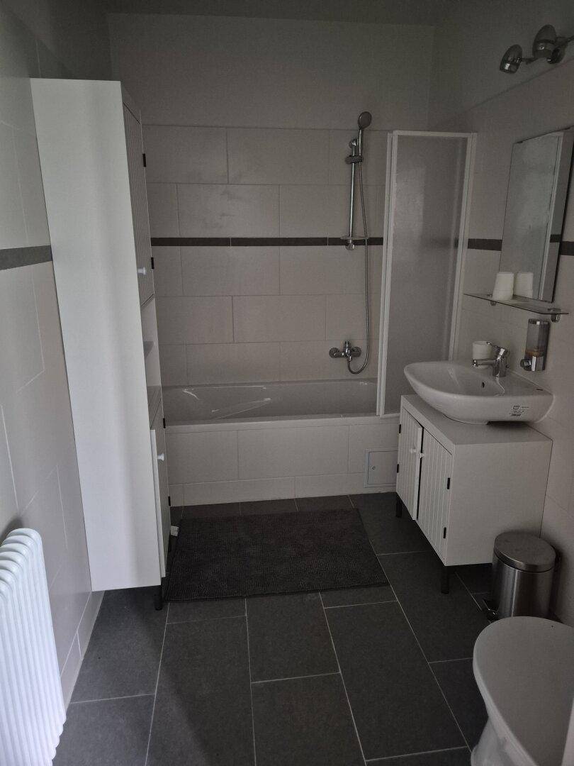 Modernes Badezimmer mit Badewanne, Duschabtrennung, Waschbecken und grauen Fliesen.